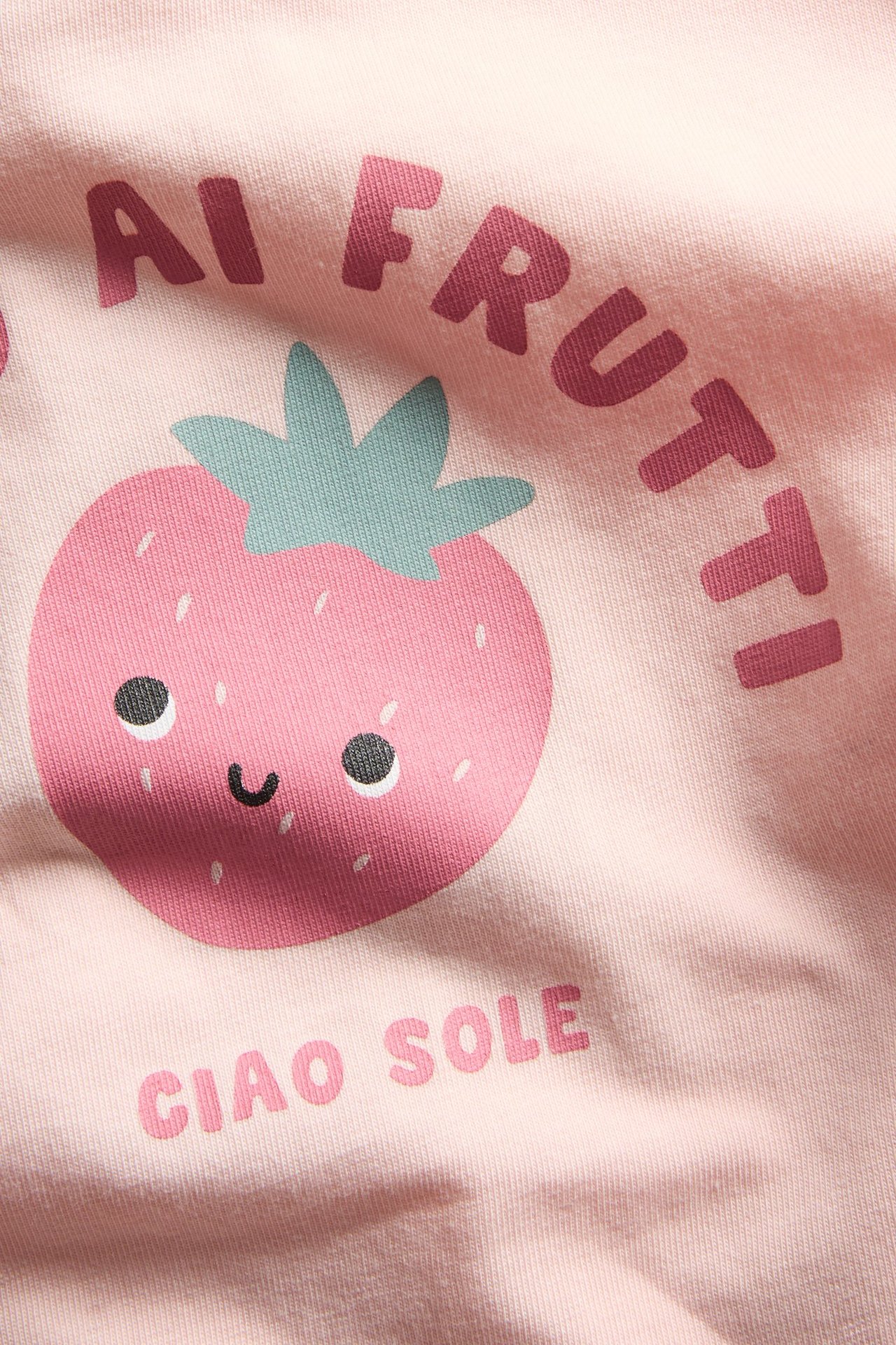 Zbliżenie na jasnoróżową koszulkę niemowlęcą z uśmiechniętym nadrukiem truskawki i tekstem z przodu.