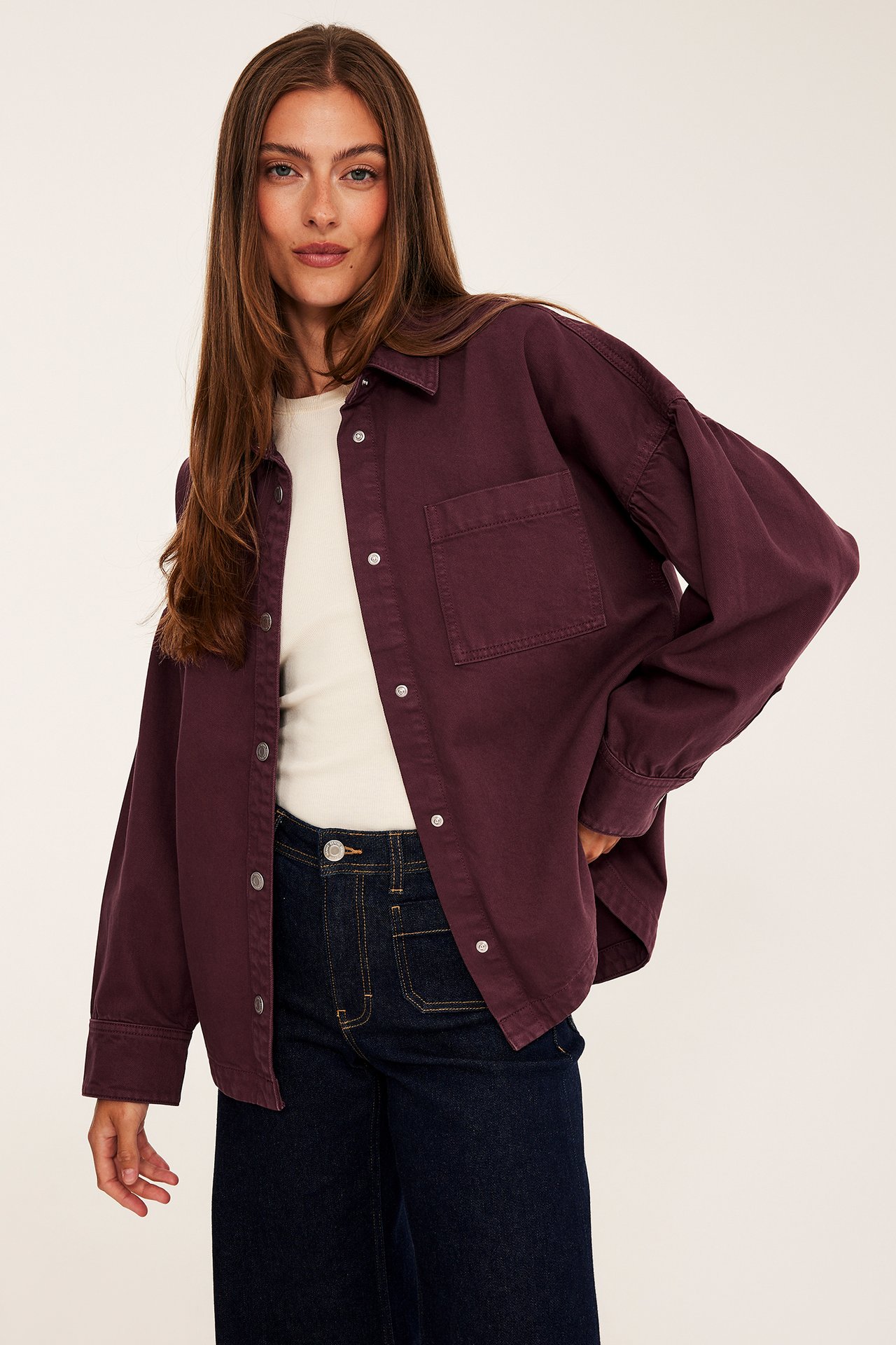 Framifrån, modell i öppen mörkröd skjortjacka över krämvit topp och mörkblå jeans.