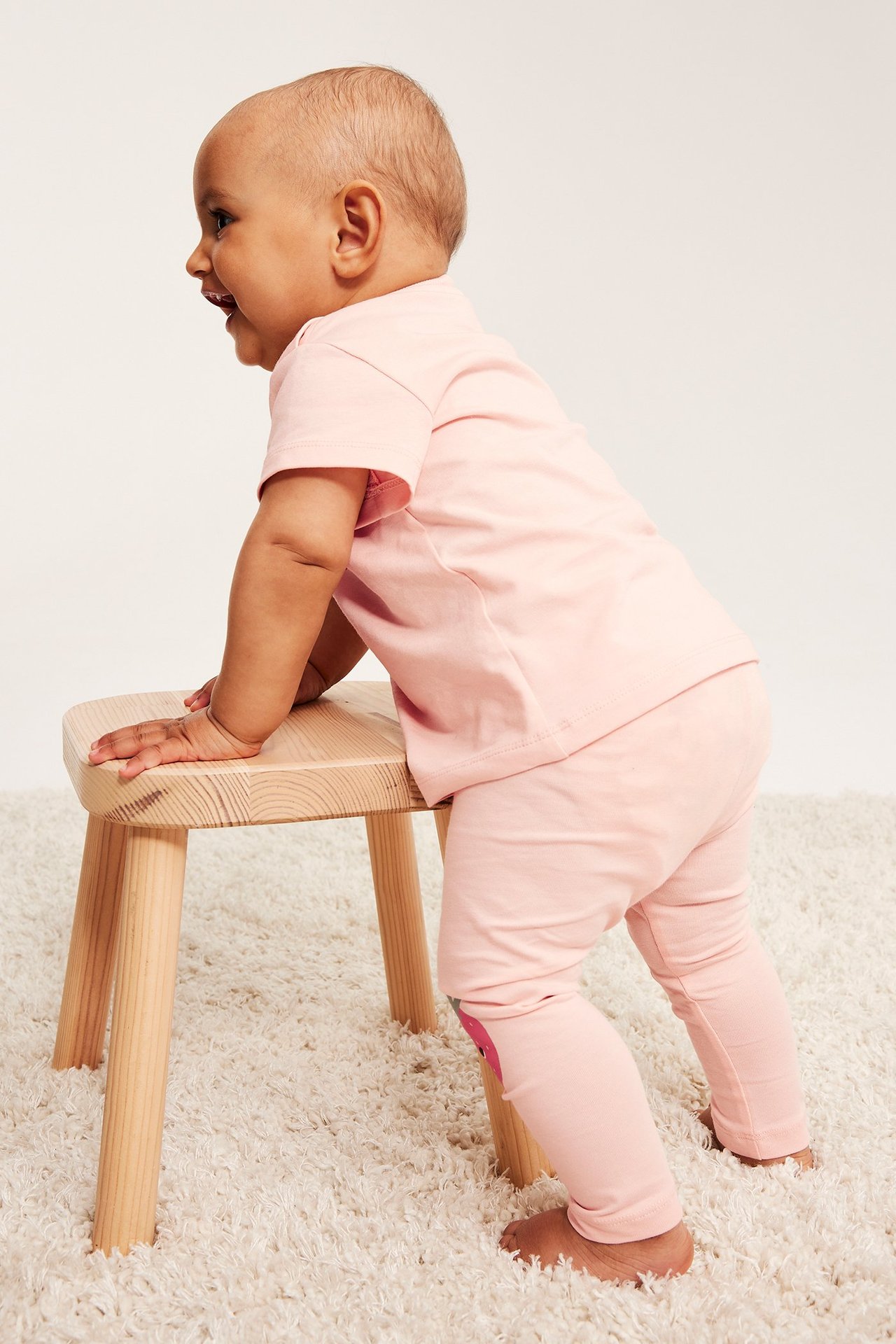 Widok z boku uśmiechniętego dziecka w jasnoróżowych bawełnianych legginsach z aplikacjami na kolanach i pasującej koszulce.