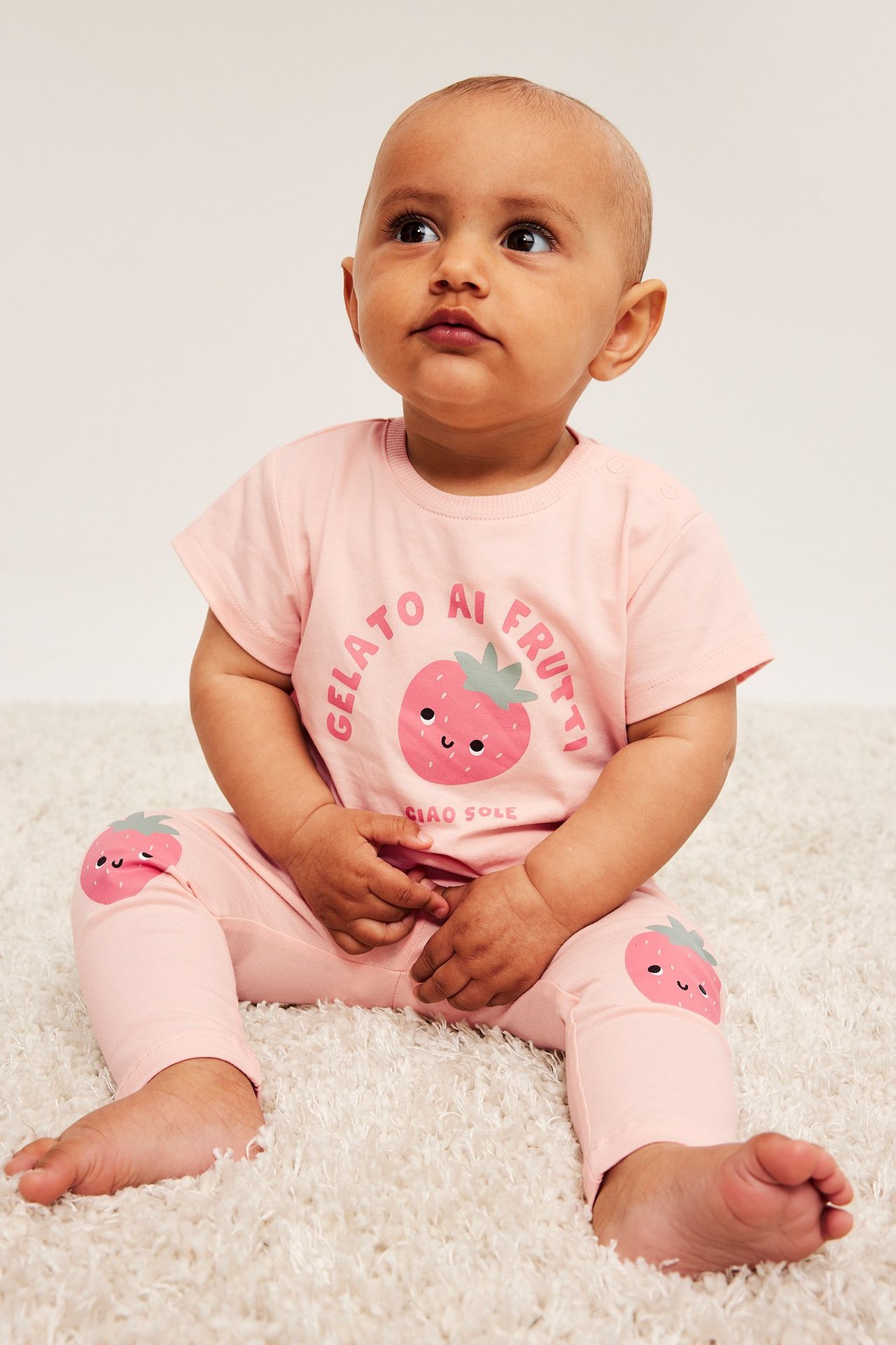 Framifrån sett, en baby sitter i ljusrosa leggings med jordgubbsapplikationer och matchande t-shirt.