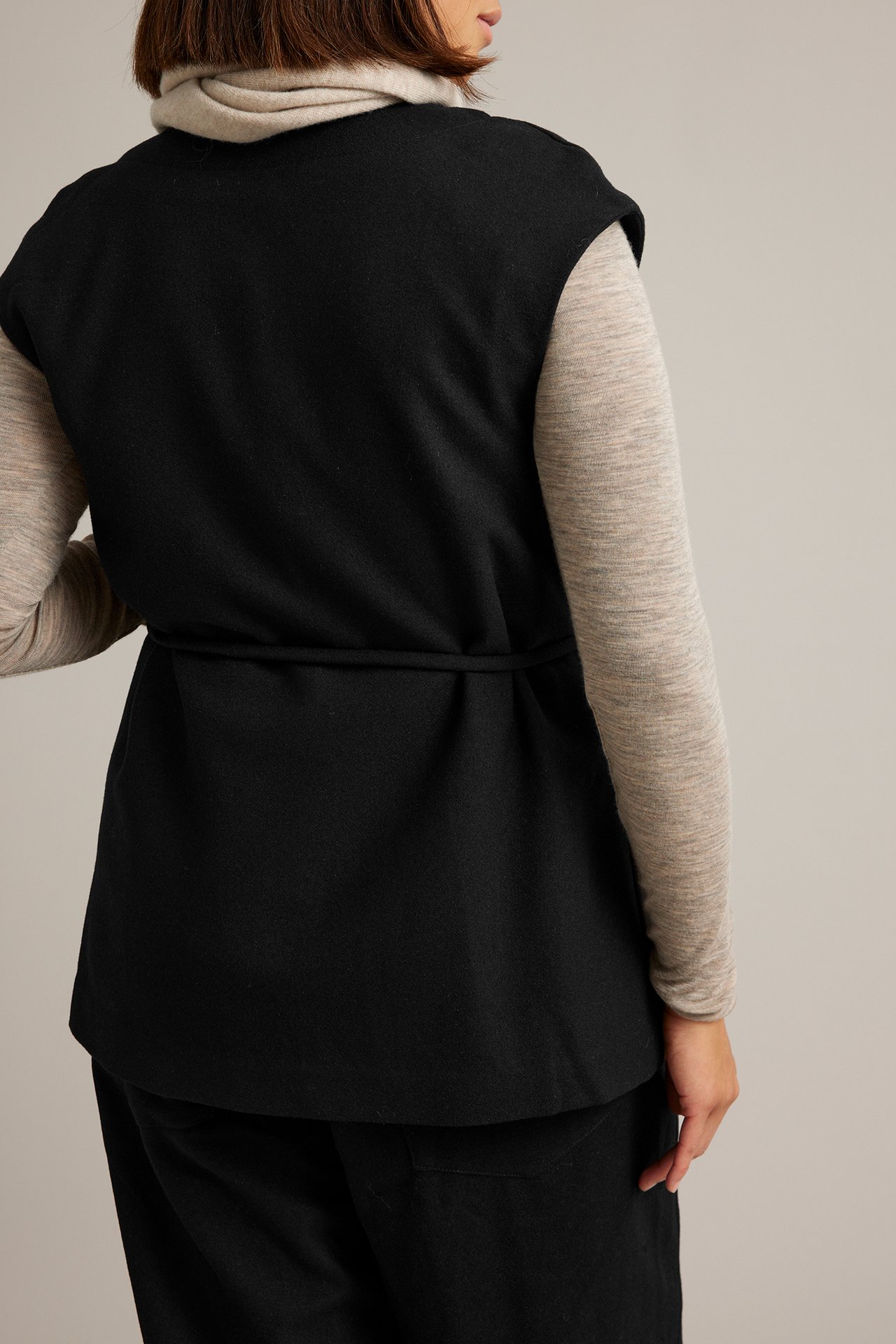 Bakfra: kvinnelig modell i svart ermeløs vest med knytebånd over beige turtleneck.