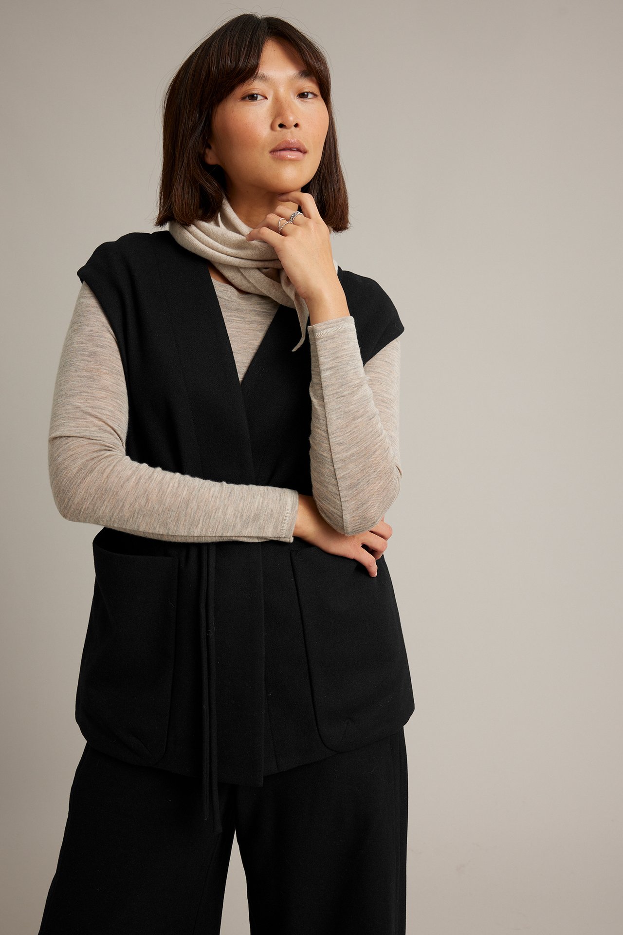 Frontbilde av kvinne i svart knytevest over beige topp og svarte bukser.
