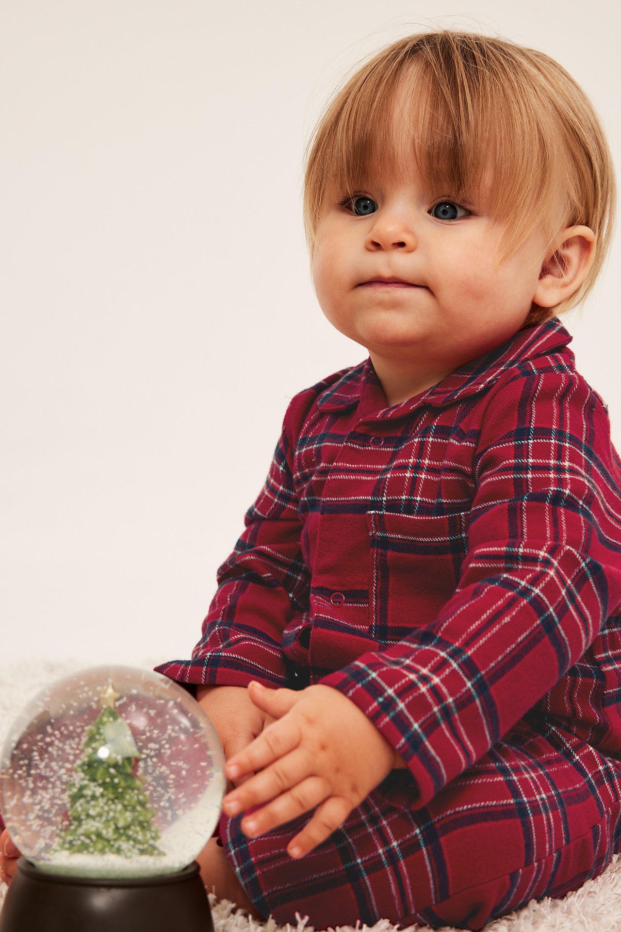 Frontbilde av baby i rød rutete pyjamas, sitter ved en snøkule.