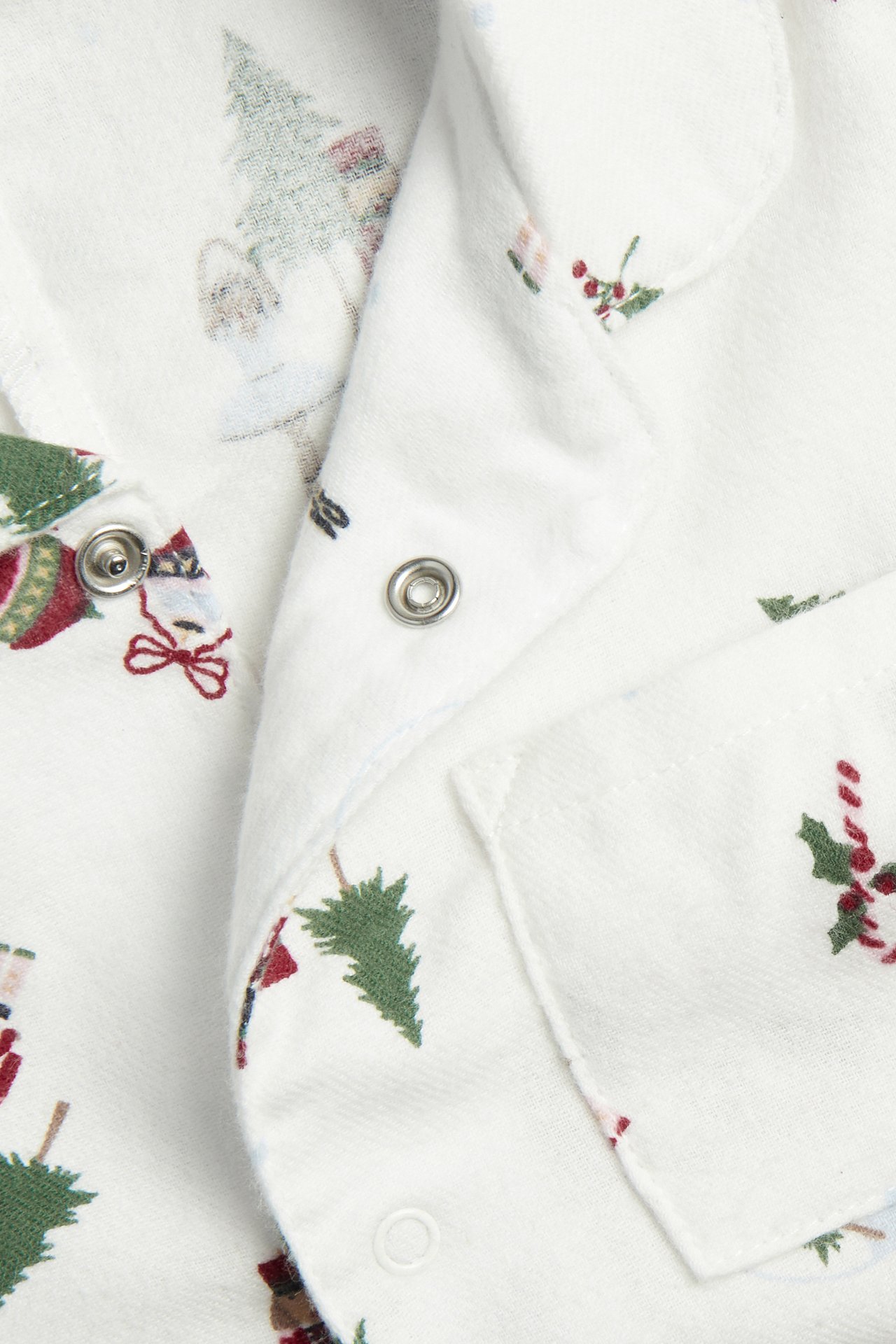 Lähikuva valkoisesta vauvan pyjamasta, jossa on joulukuusi-, karkkikeppi- ja lumiukkokuvio, hopeisilla neppareilla.