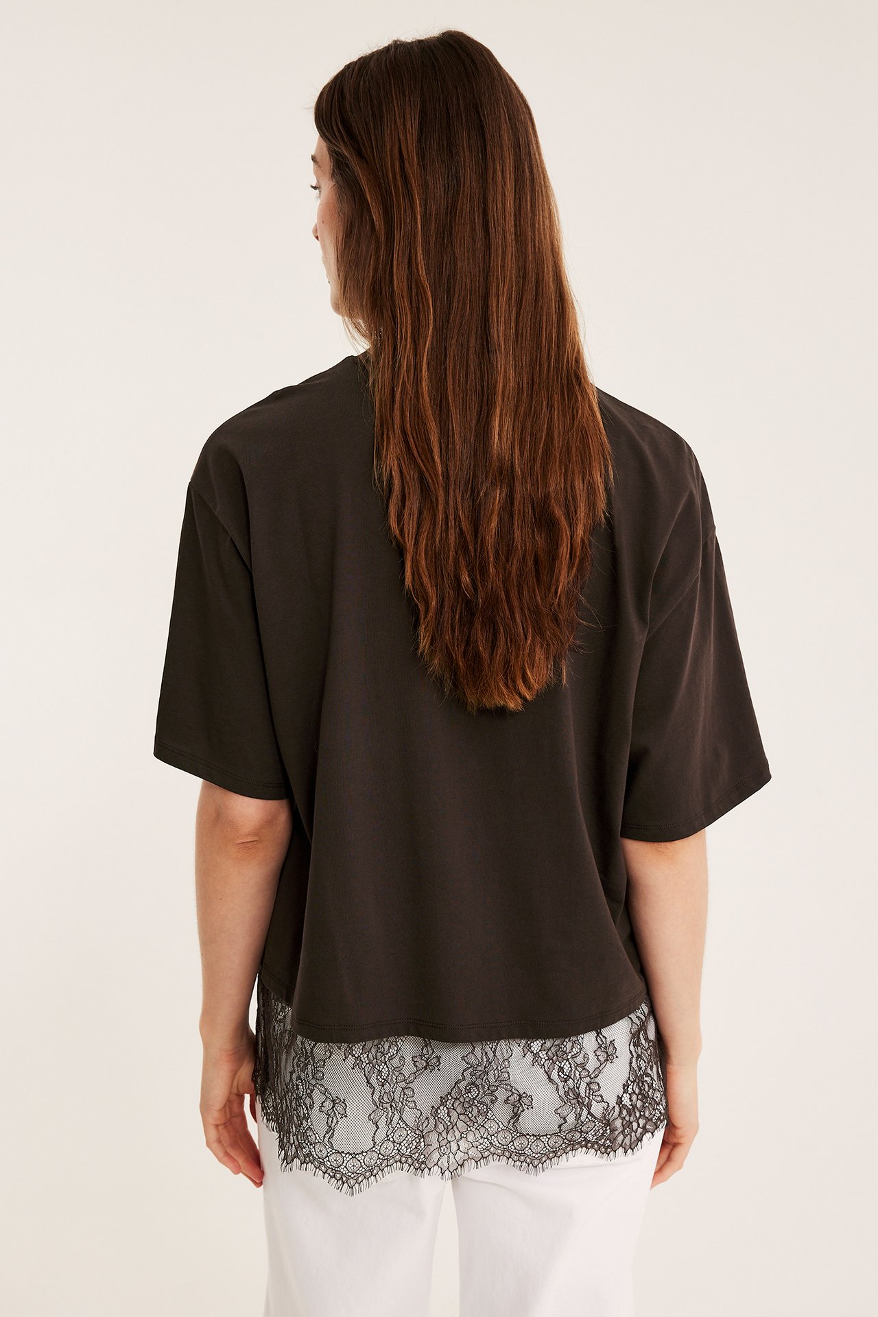Ryggvy av en kvinna i en lös mörkbrun t-shirt med svart spetskant över vita byxor.