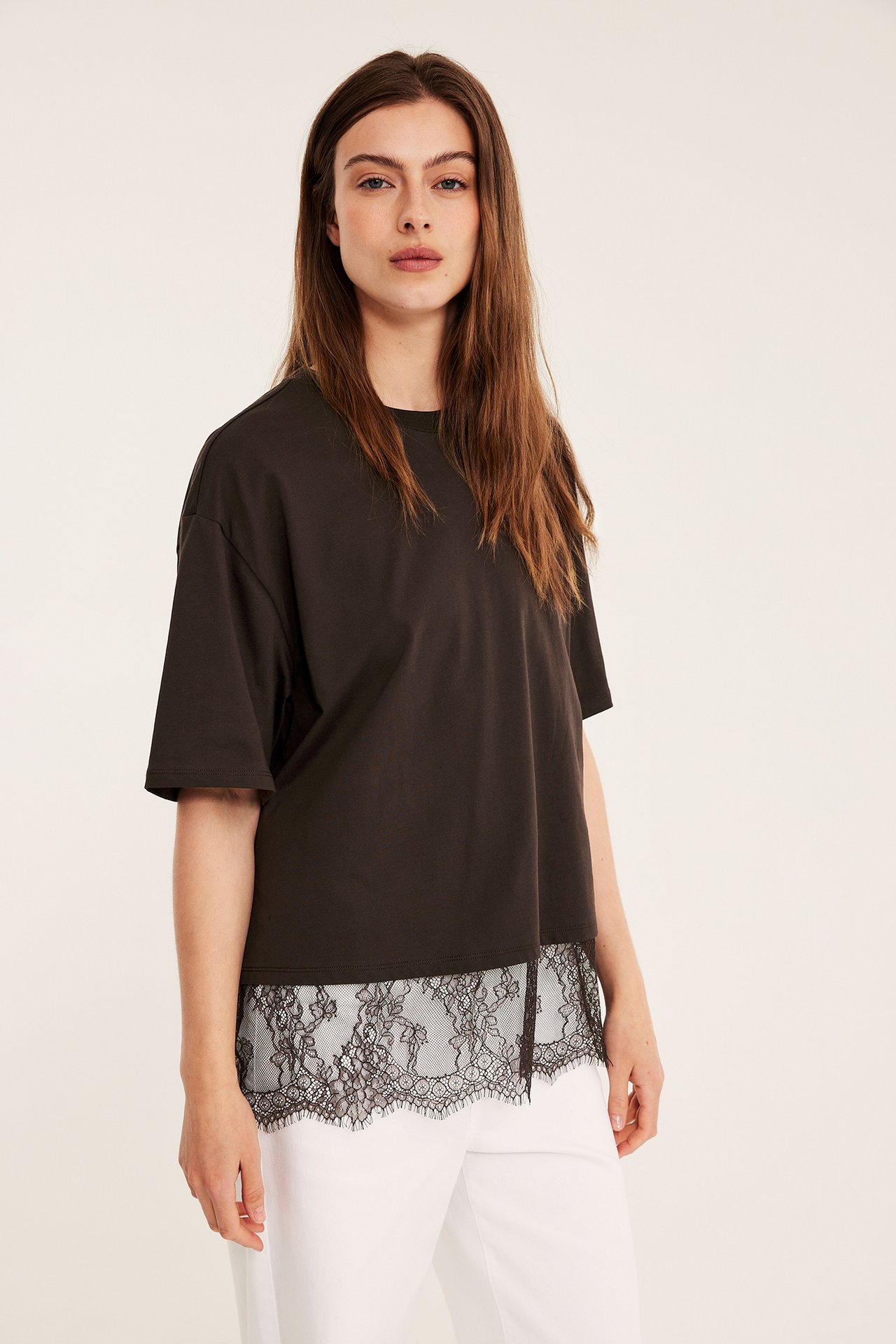 Framifrån, kvinna i lös mörkbrun t-shirt med svart spetskant, stylad med vita byxor.