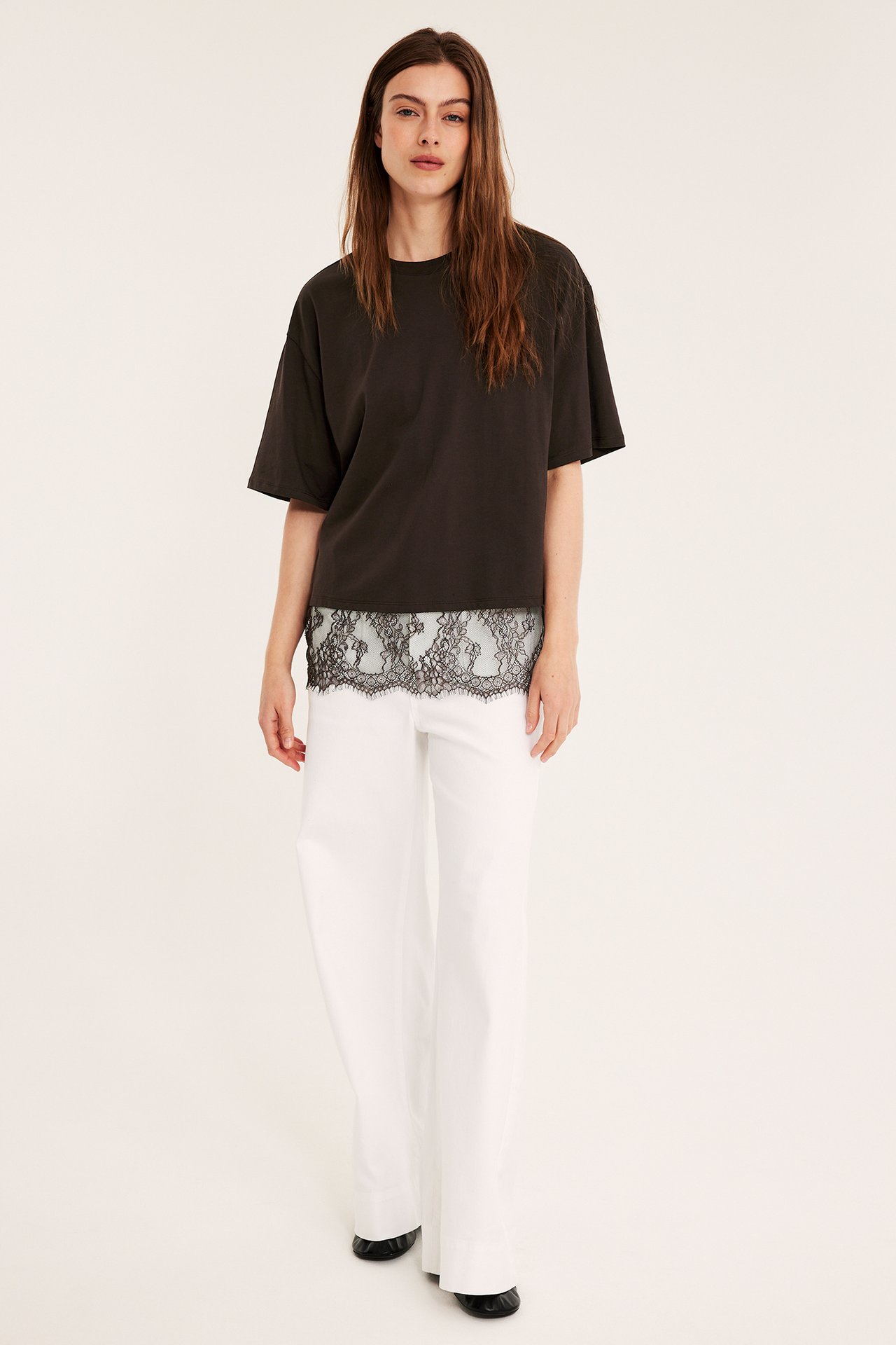 Framifrån: Kvinna i lös mörkbrun t-shirt med svart spets, vita vida byxor, svarta platta skor.