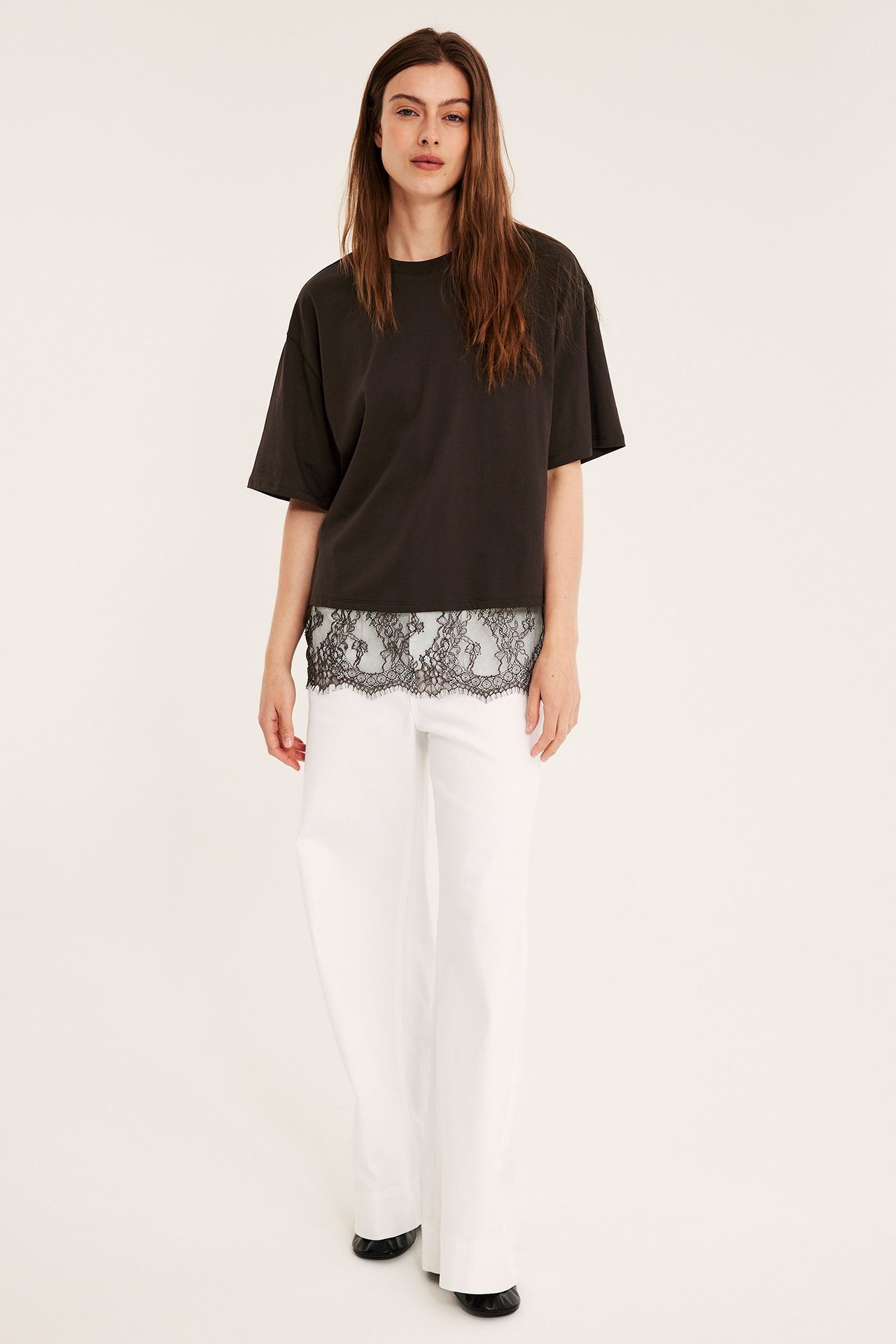 Edestäpäin: Nainen löysässä tummanruskeassa t-paidassa mustalla pitsillä, valkoisissa leveissä housuissa, mustissa kengissä.