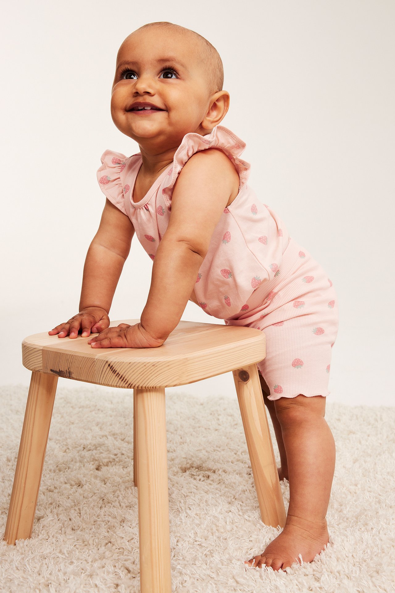 Widok z przodu: niemowlę w jasnoróżowych, prążkowanych szortach rowerowych w truskawki i pasującym topie z falbankami.