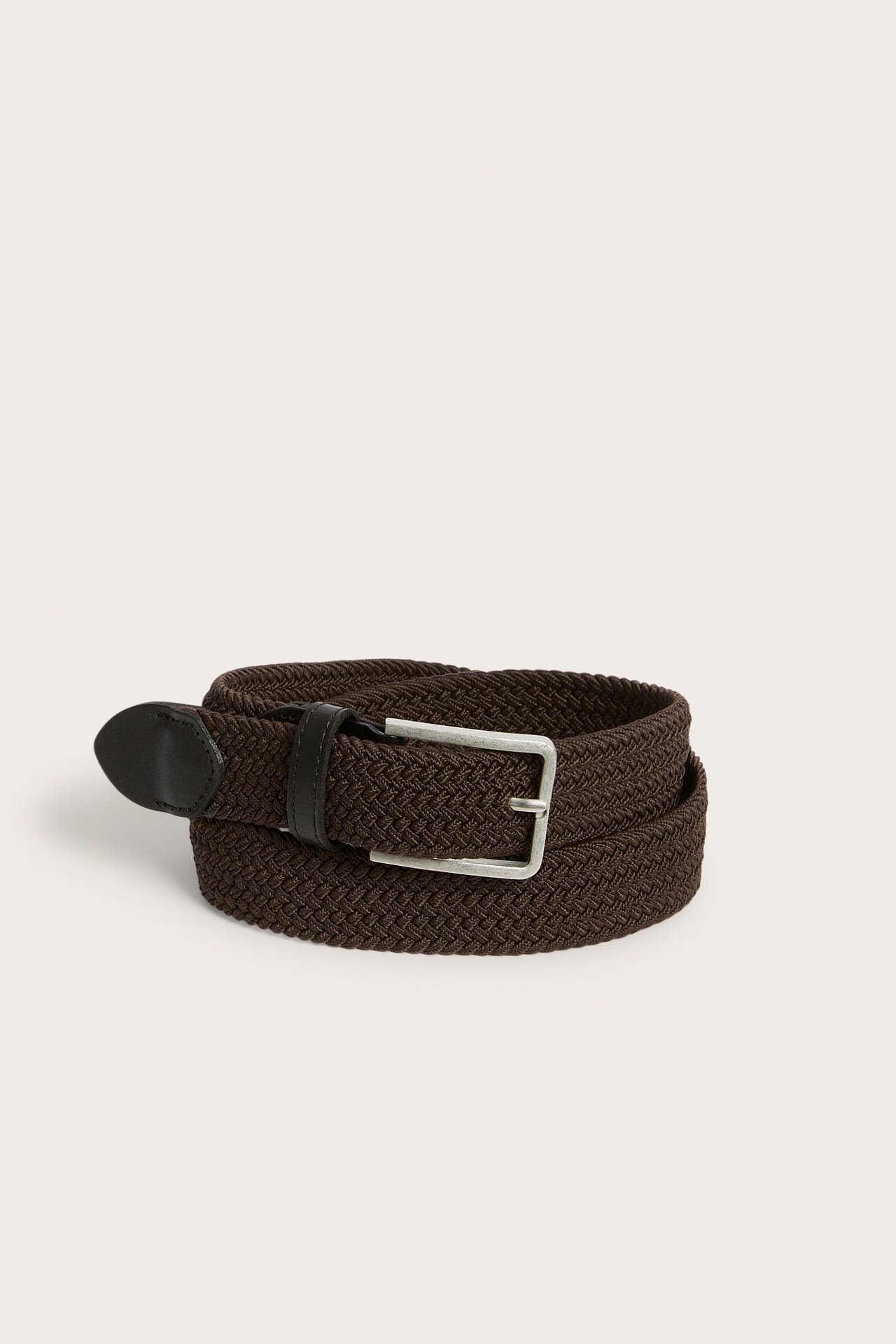 Närbild av ett lindat mörkbrunt flätat elastiskt bälte med svart lädertipp och silverspänne.