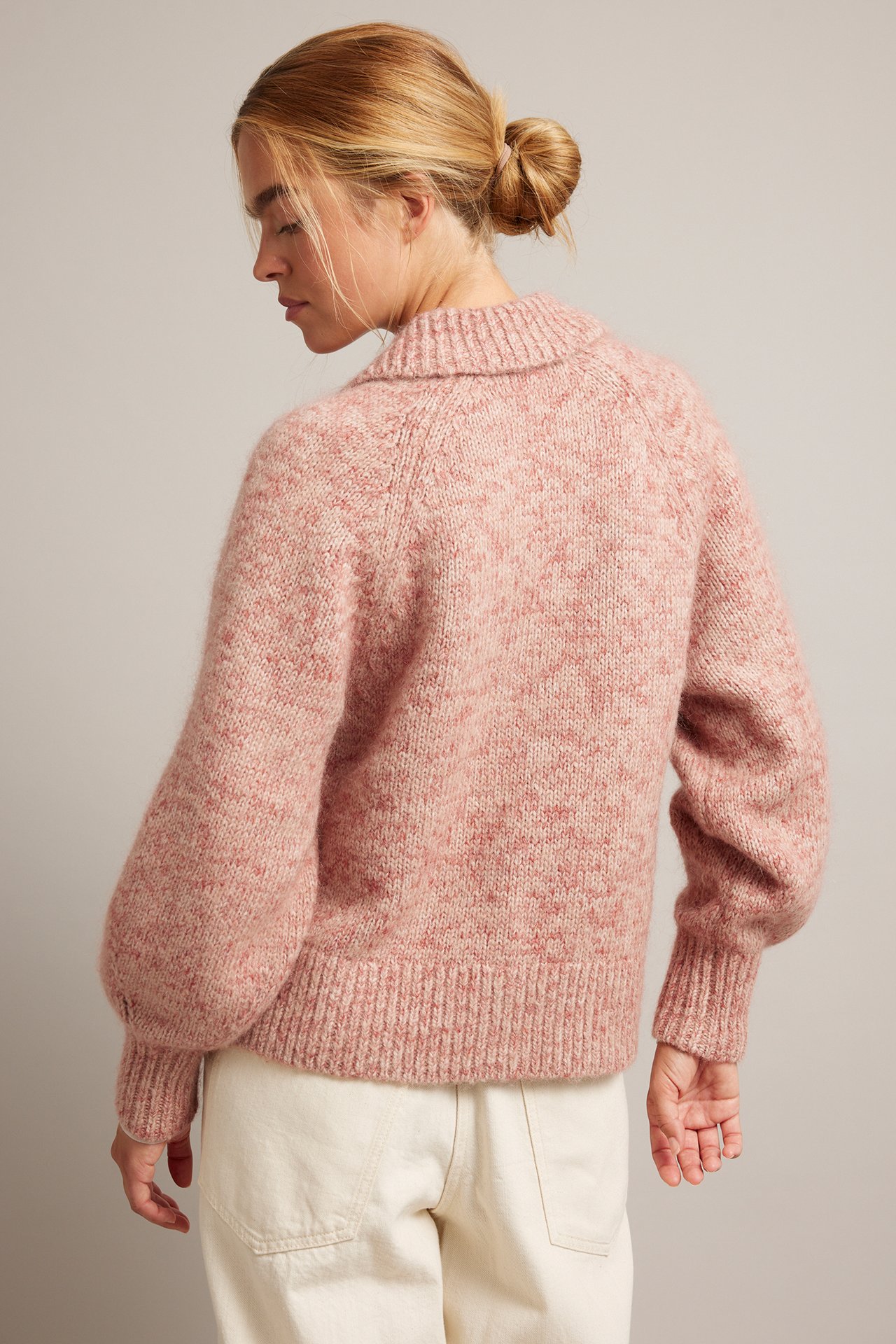 Bakifrån kvinnlig modell i rosa melerad stickad tröja med krage, ballongärmar och benvita byxor.