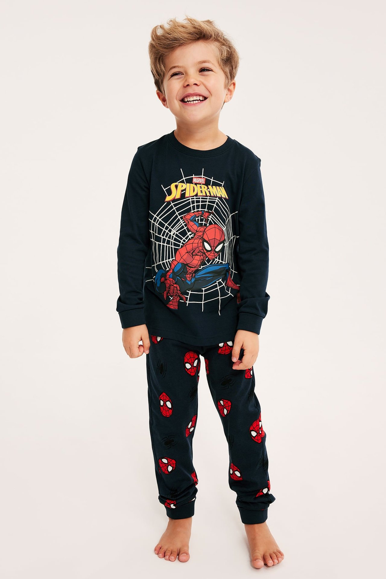 Forfra: Smilende barn i marineblå langermet Spiderman-pyjamas med trykte masker.