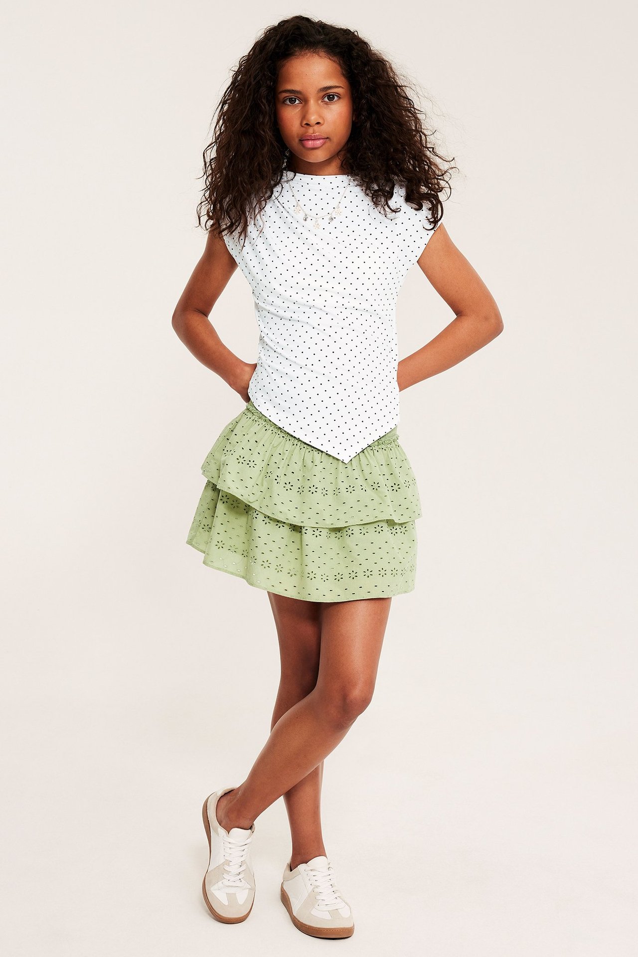 Frontvisning: Barn i stram hvit prikkete asymmetrisk topp, lysegrønt skjørt med volanger og hvite joggesko.