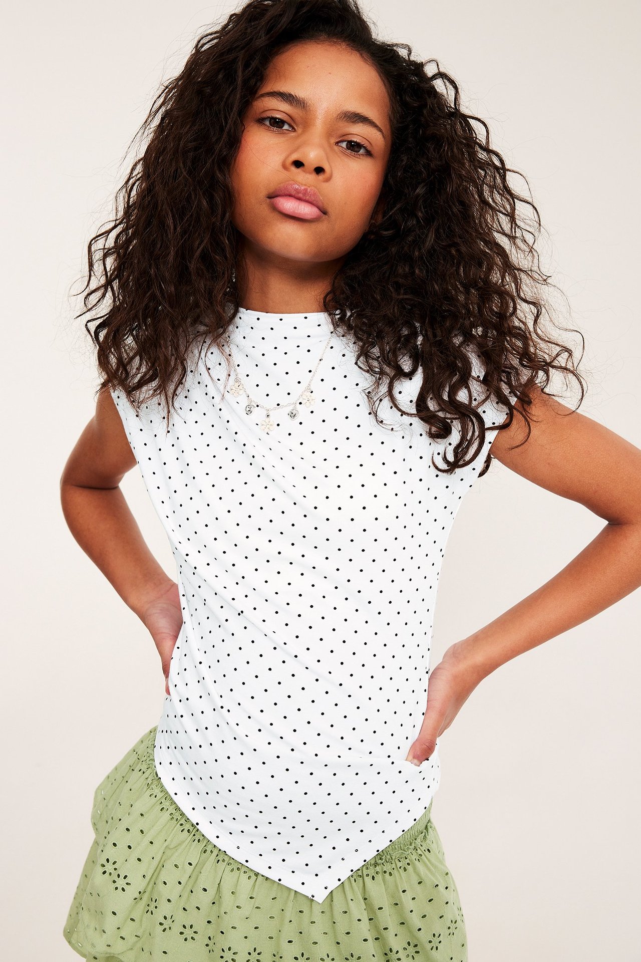 Widok z przodu dziecka w białym topie w kropki z asymetrycznym dołem i zielonej haftowanej spódnicy.
