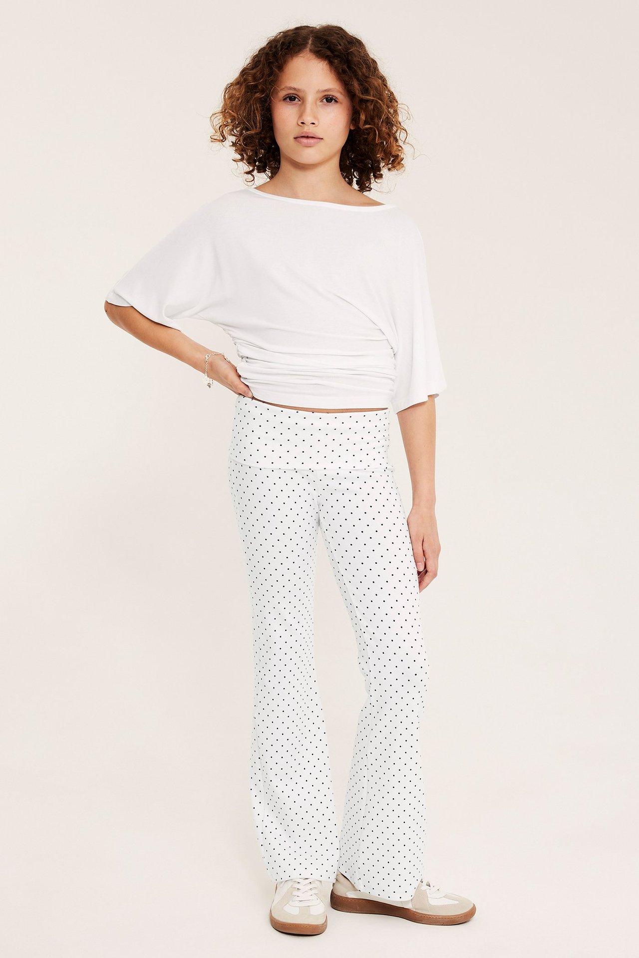 Lapsi edestä valkoisissa pilkullisissa levenevissä leggingseissä, valkoisessa topissa ja beigeissä lenkkareissa.