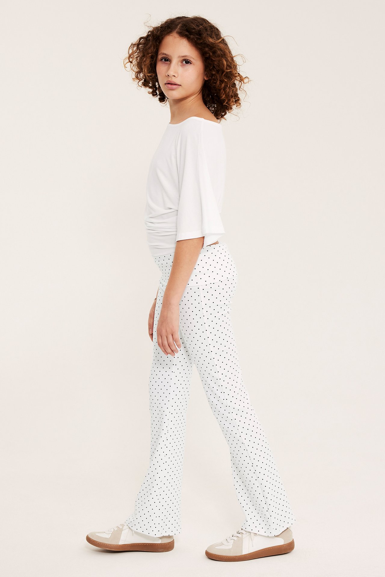 Sidovy av ett barn som bär vita prickiga utsvängda yogabyxor, en vit långärmad topp och sneakers.