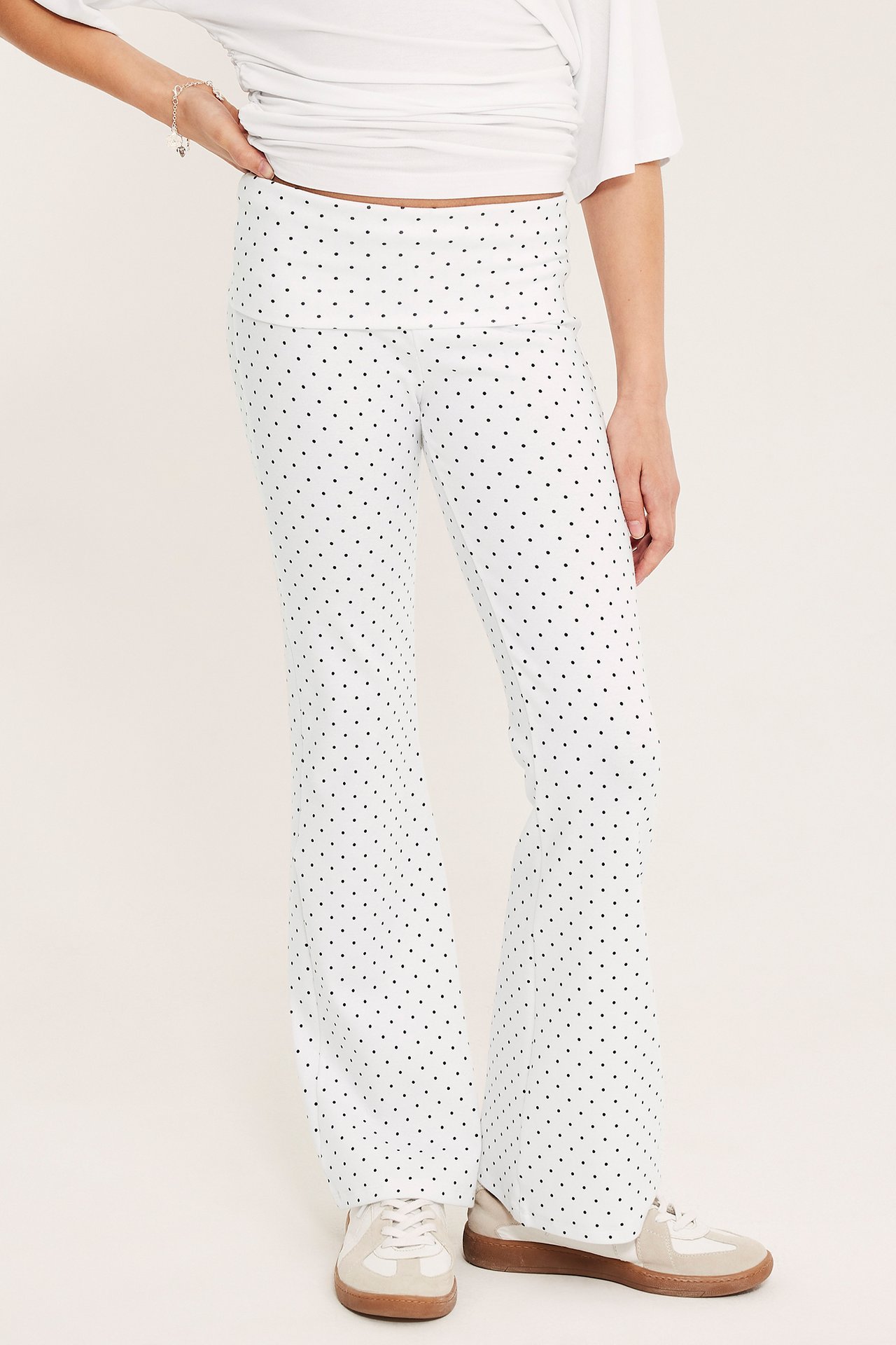 Widok z przodu: Dziecko w białych, rozszerzanych legginsach w kropki, zakładana talia, biały top, beżowe trampki.