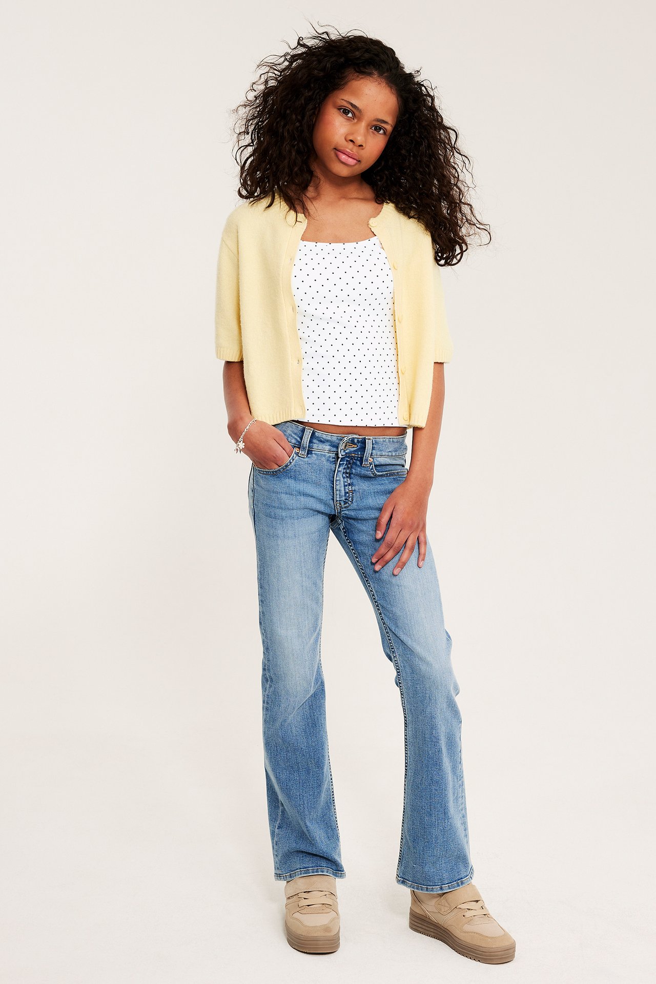 Dziecko z przodu: biały top w kropki, żółty kardigan, niebieskie dżinsy, beżowe trampki.