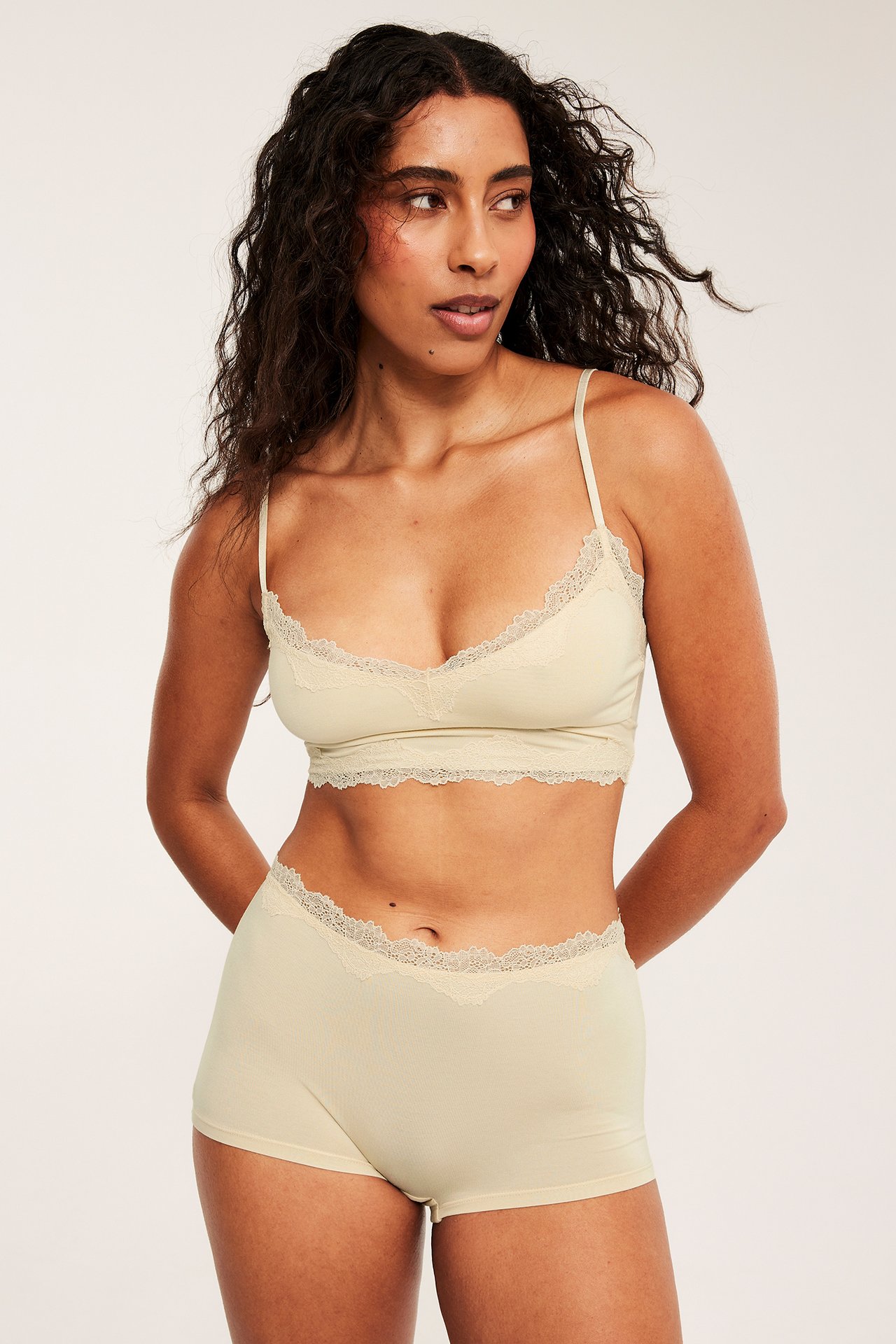 Framifrån: kvinna i beige modal spetsbralette med matchande spetskantade boy shorts.