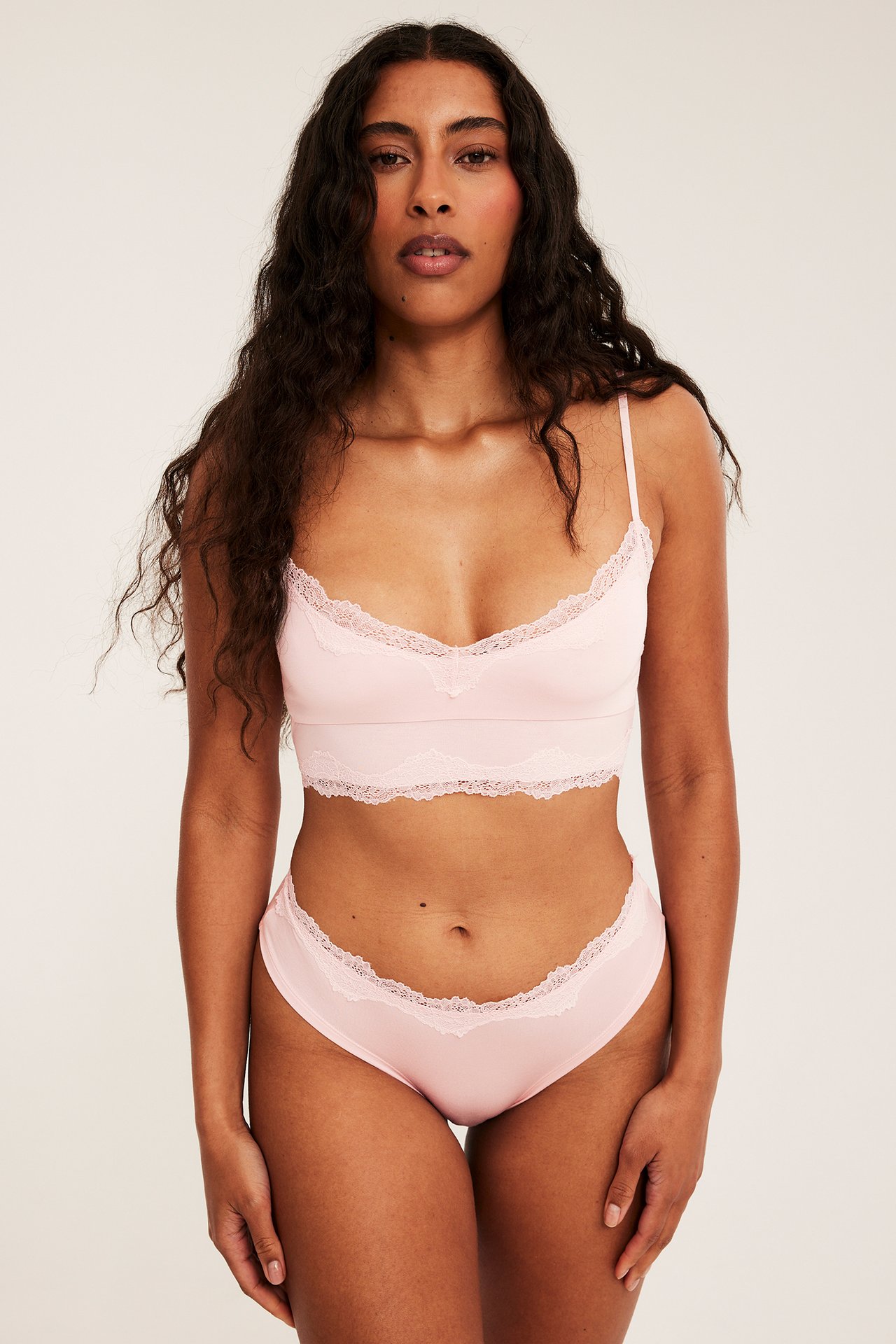 Framifrån, kvinna i ljusrosa cheeky trosa av modal med spetskant, samt matchande bralette.