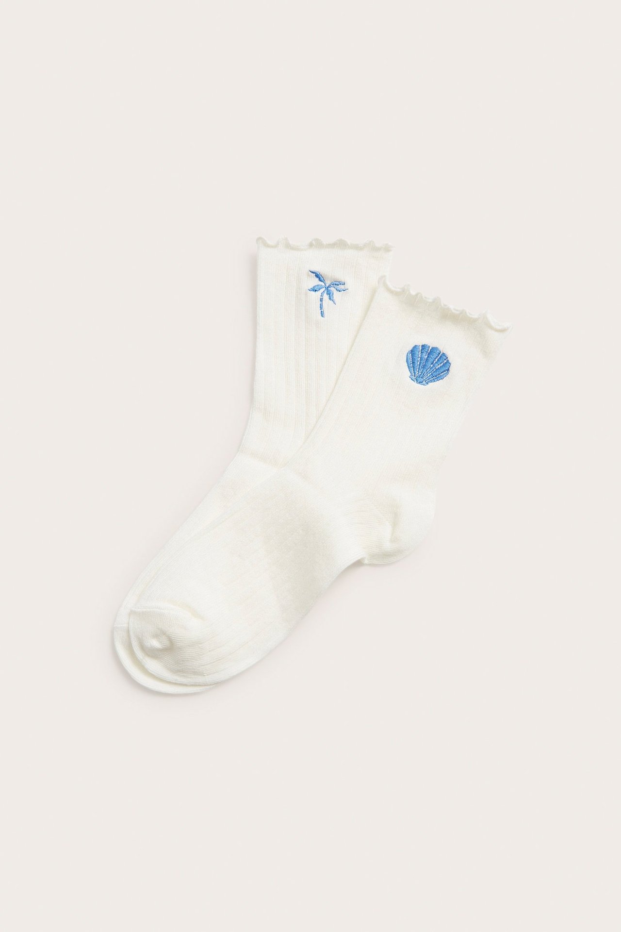 Widok z przodu: dwie jasnobeżowe prążkowane skarpetki z falbanką, haftowane w niebieską palmę i muszelkę.