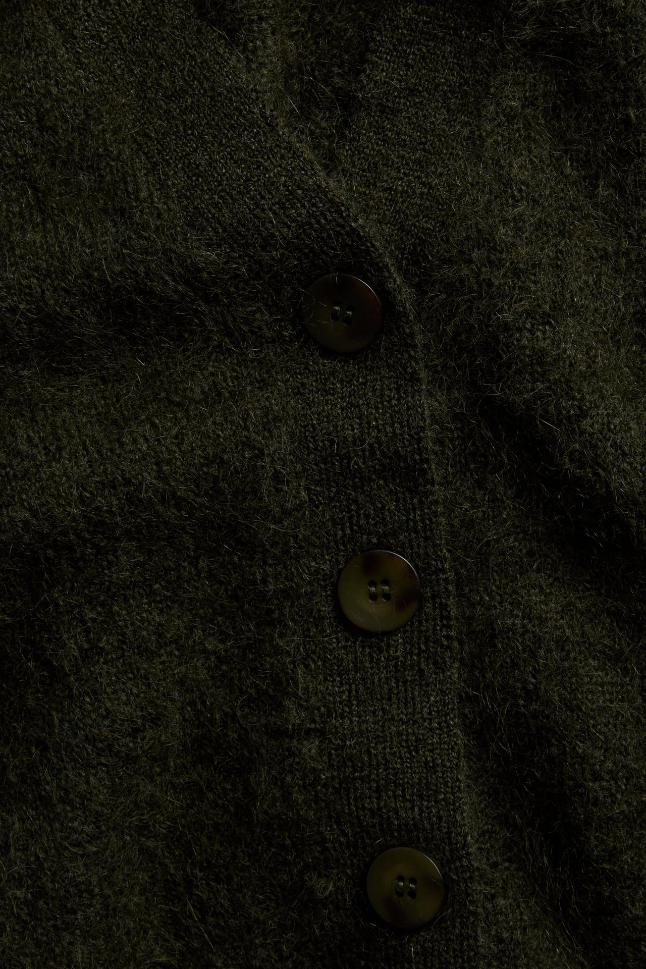 Nærbilde av en mørkegrønn lodden strikket cardigan med tre mørke knapper.