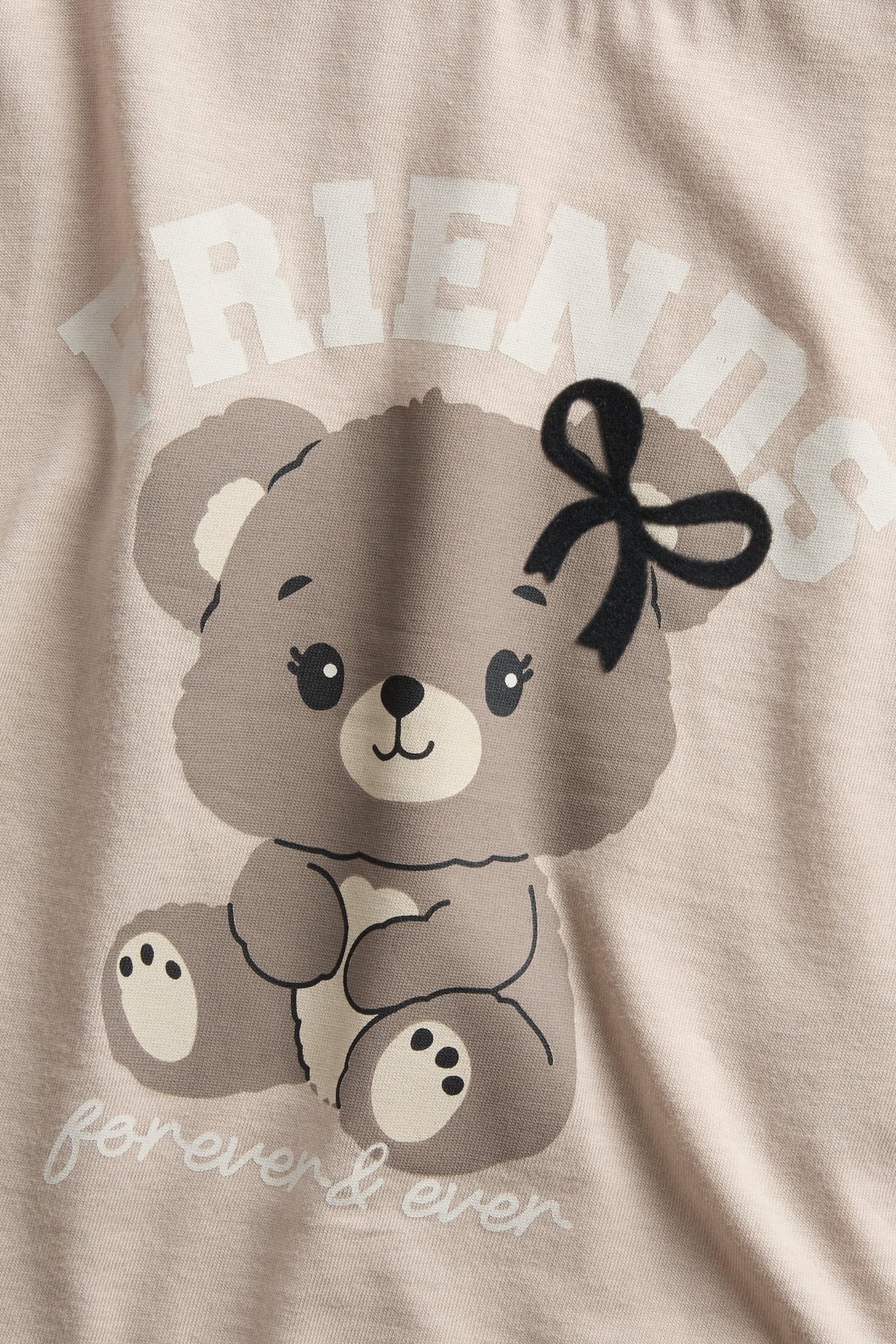 Närbild av ljusbeige textil med 'FRIENDS forever & ever' nallebjörnstryck.