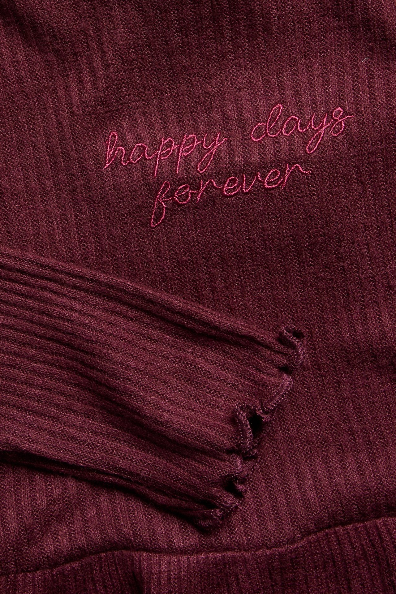 Närbild på en vinröd ribbad topp med broderad rosa text 'happy days forever' och fransad manschett.