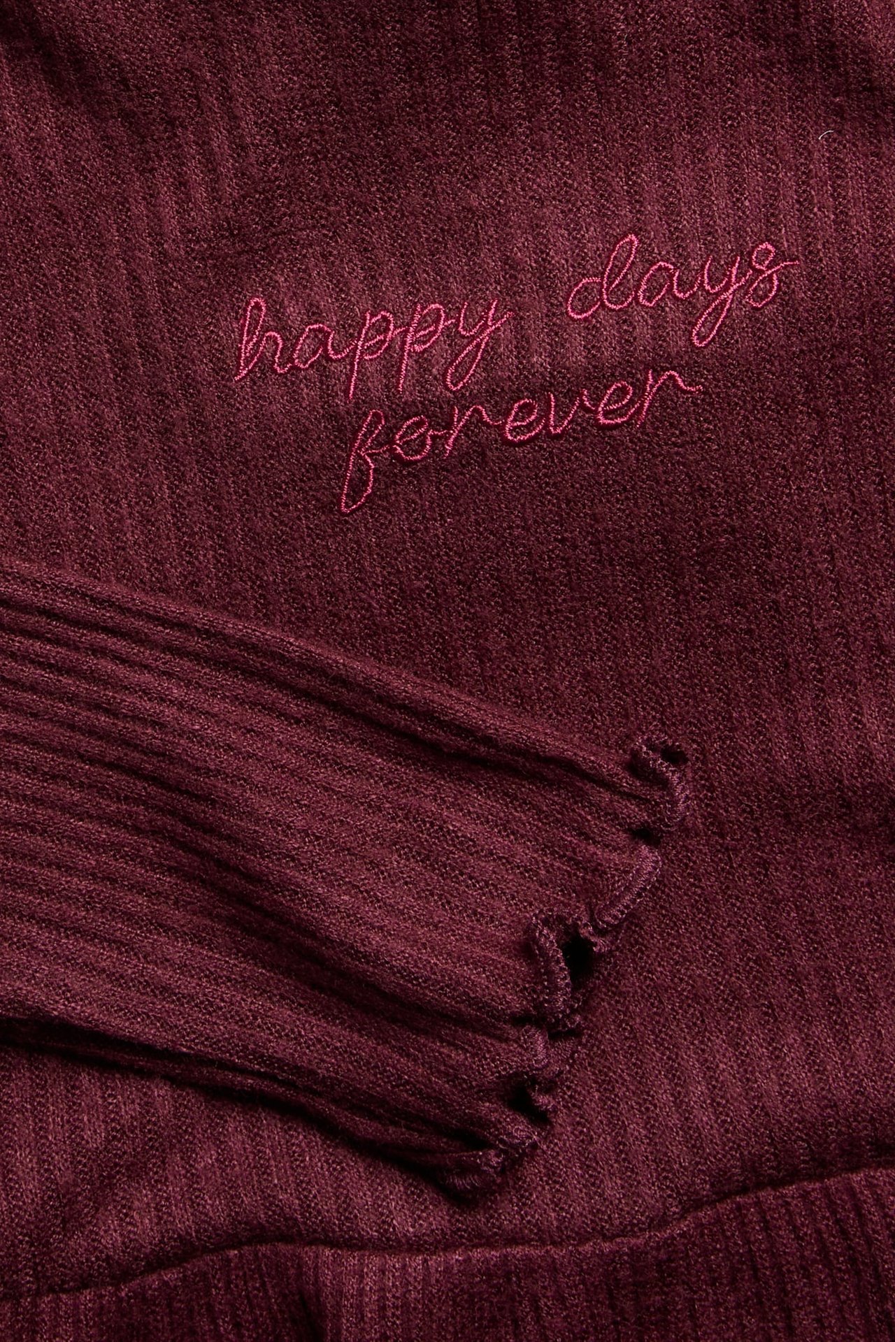 Lähikuva viininpunaisesta ribbipaidasta, jossa on brodeerattu pinkki teksti 'happy days forever' ja hapsuinen ranneke.