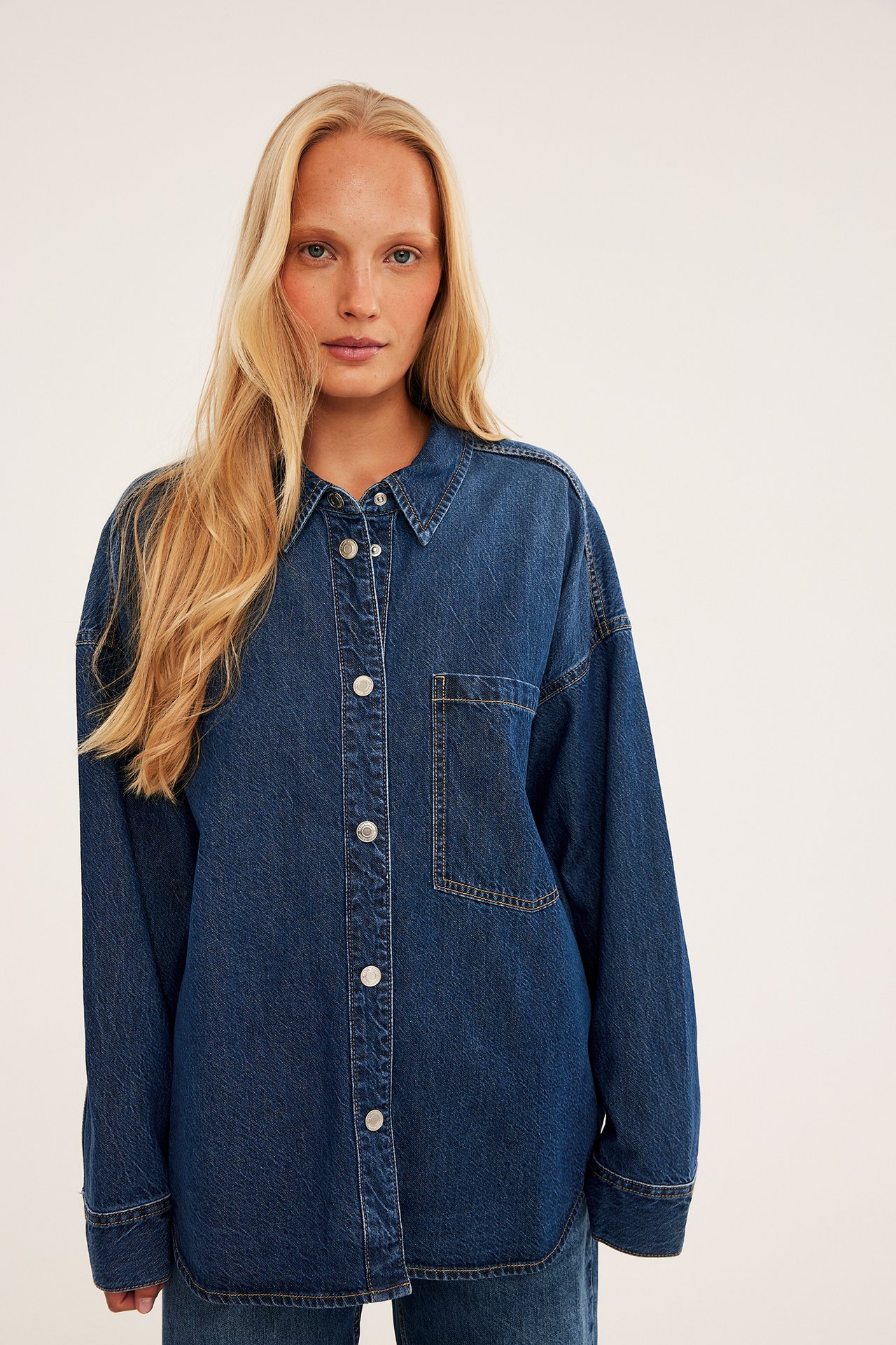 Front av kvinne i en oversized mørkblå denimskjorte med trykknapper og brystlomme.