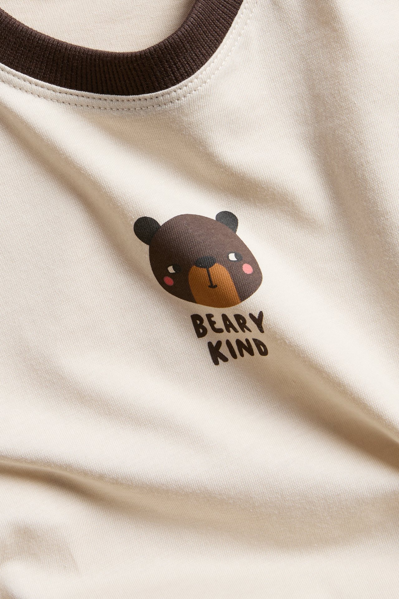 Zbliżenie jasnobeżowego dziecięcego topu z brązowym ściągaczem przy szyi i nadrukiem misia 'BEARY KIND'.