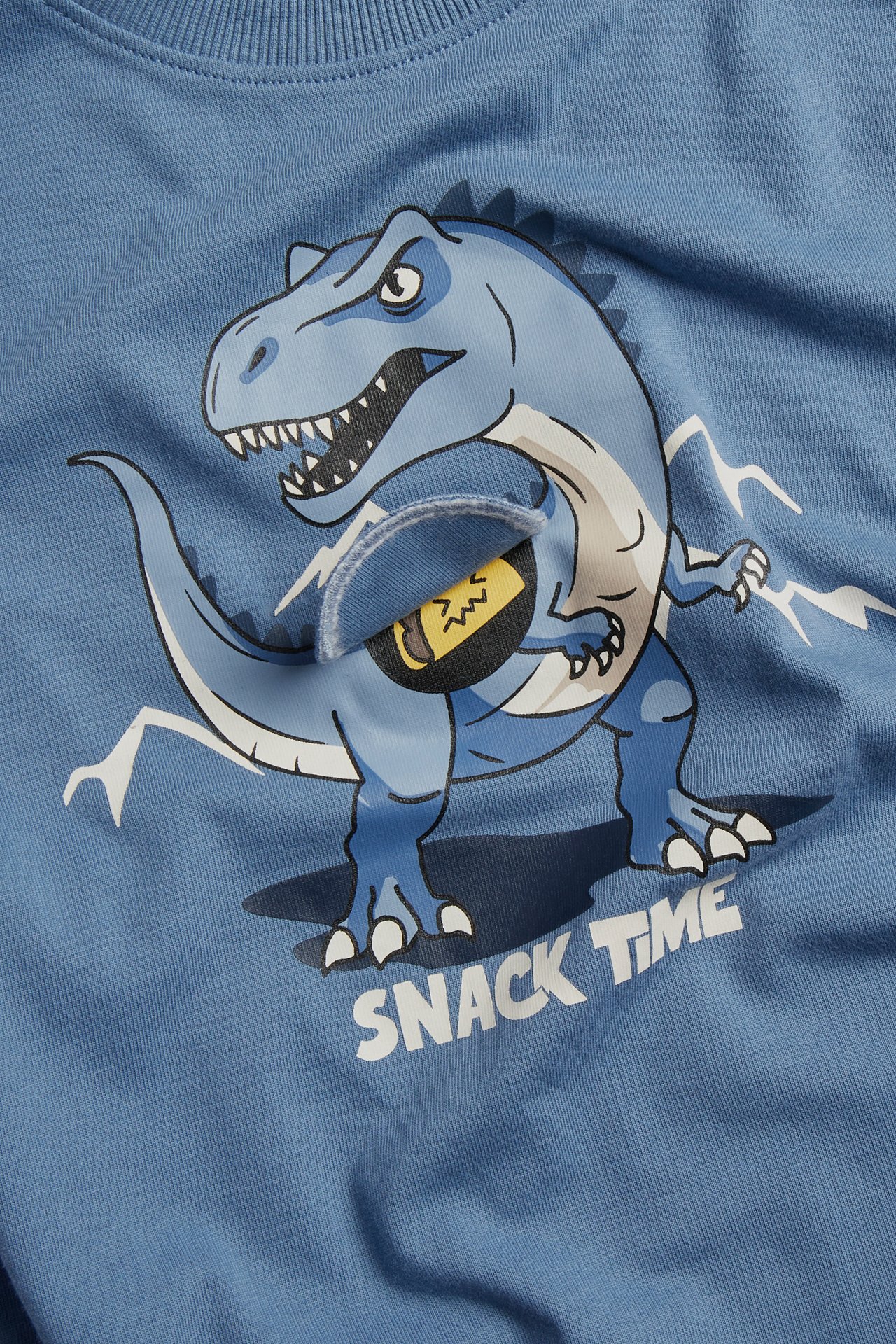 Närbild på en blå barntröja med dinosauriegrafik, en snacklucka och texten 'SNACK TIME'.