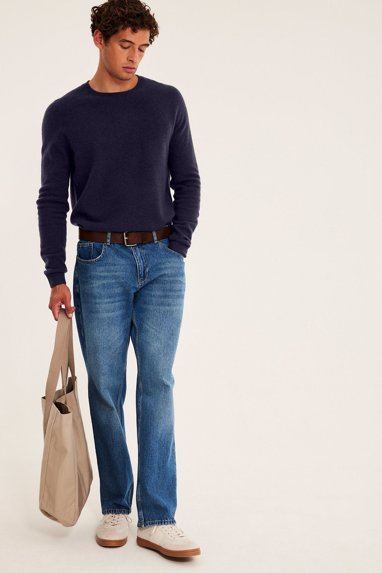 Męski model w granatowym swetrze, niebieskich jeansach, brązowym pasku, jasnych sneakersach, trzyma beżową torbę.