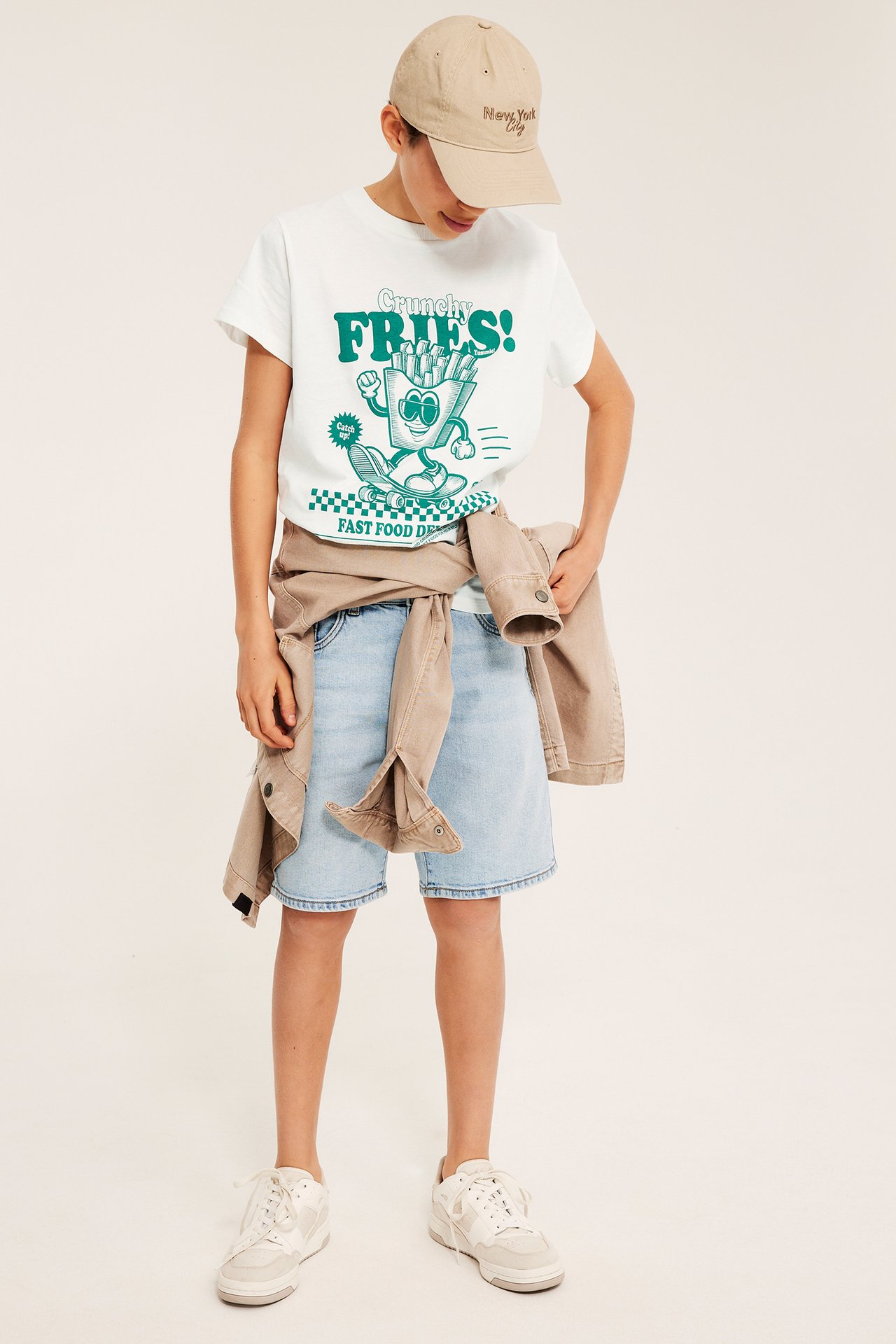 Lapsi offwhite T-paidassa 'Crunchy Fries' -painatuksella, vaaleansiniset shortsit, beige takki, lippis ja tennarit.