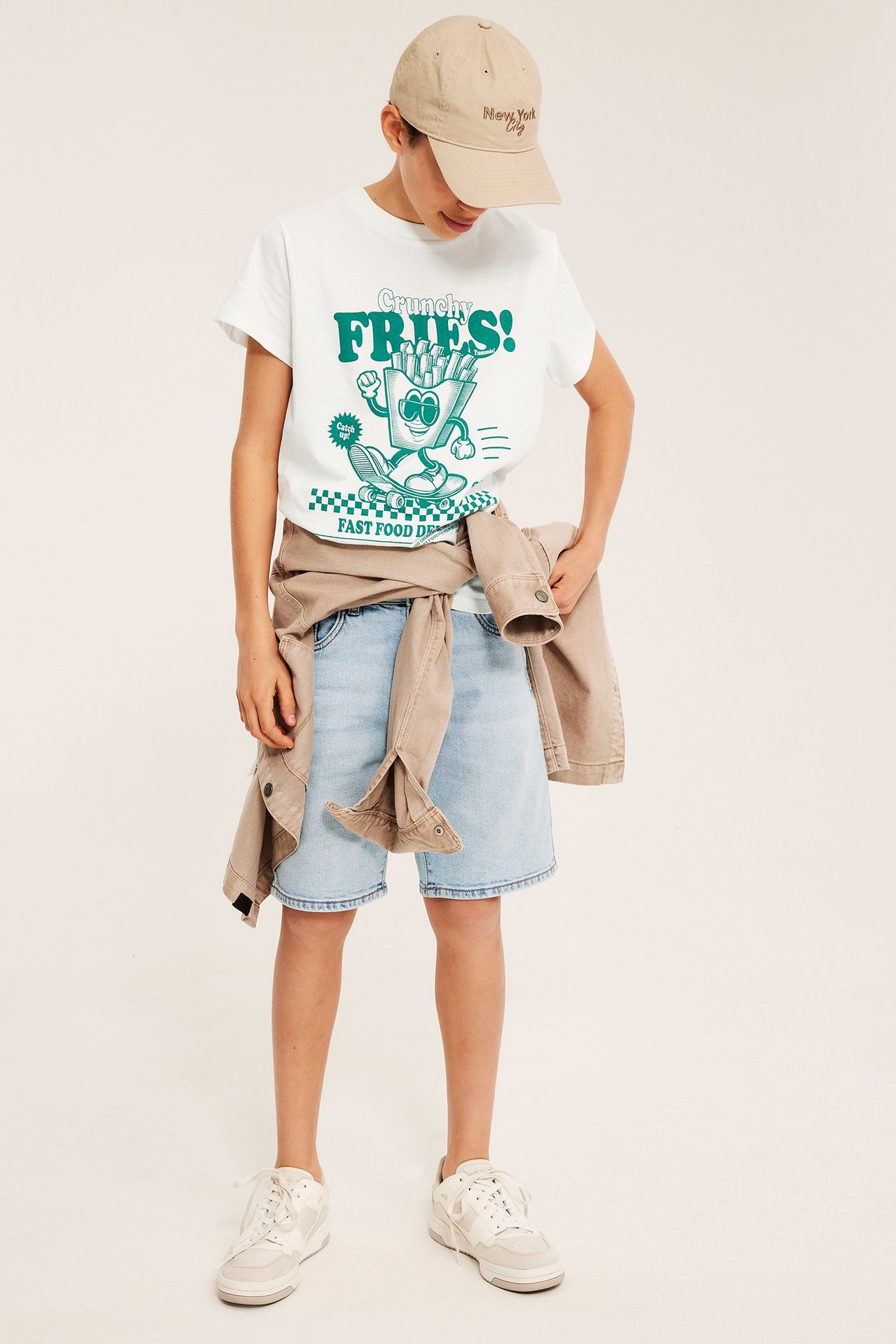 Barn i offwhite T-shirt med tryck 'Crunchy Fries', ljusblå shorts, beige jacka, keps och sneakers.