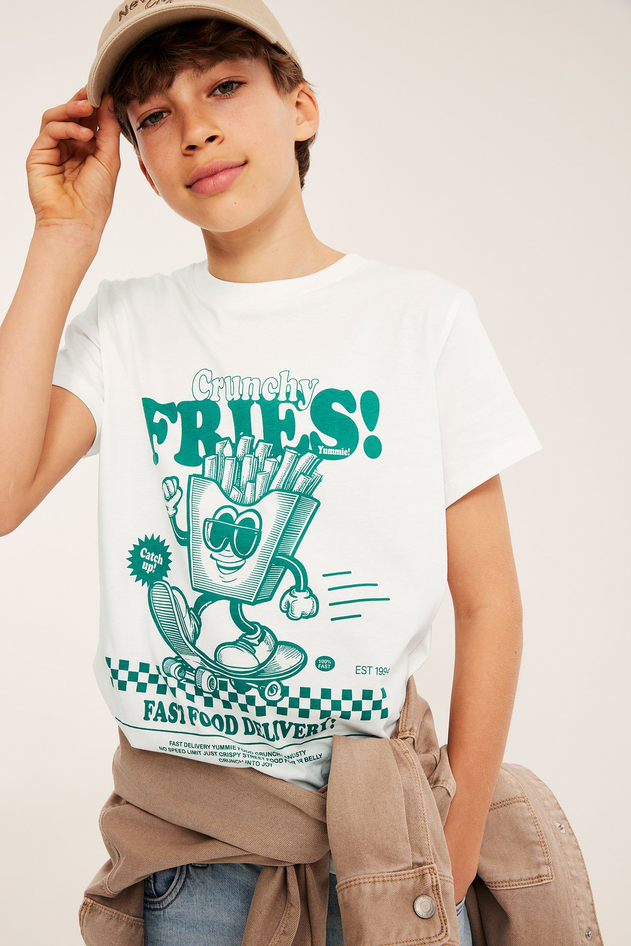 Frontvy: Ett barn i offwhite bomulls-T-shirt med grönt pommes-tryck, beige keps och jeans.