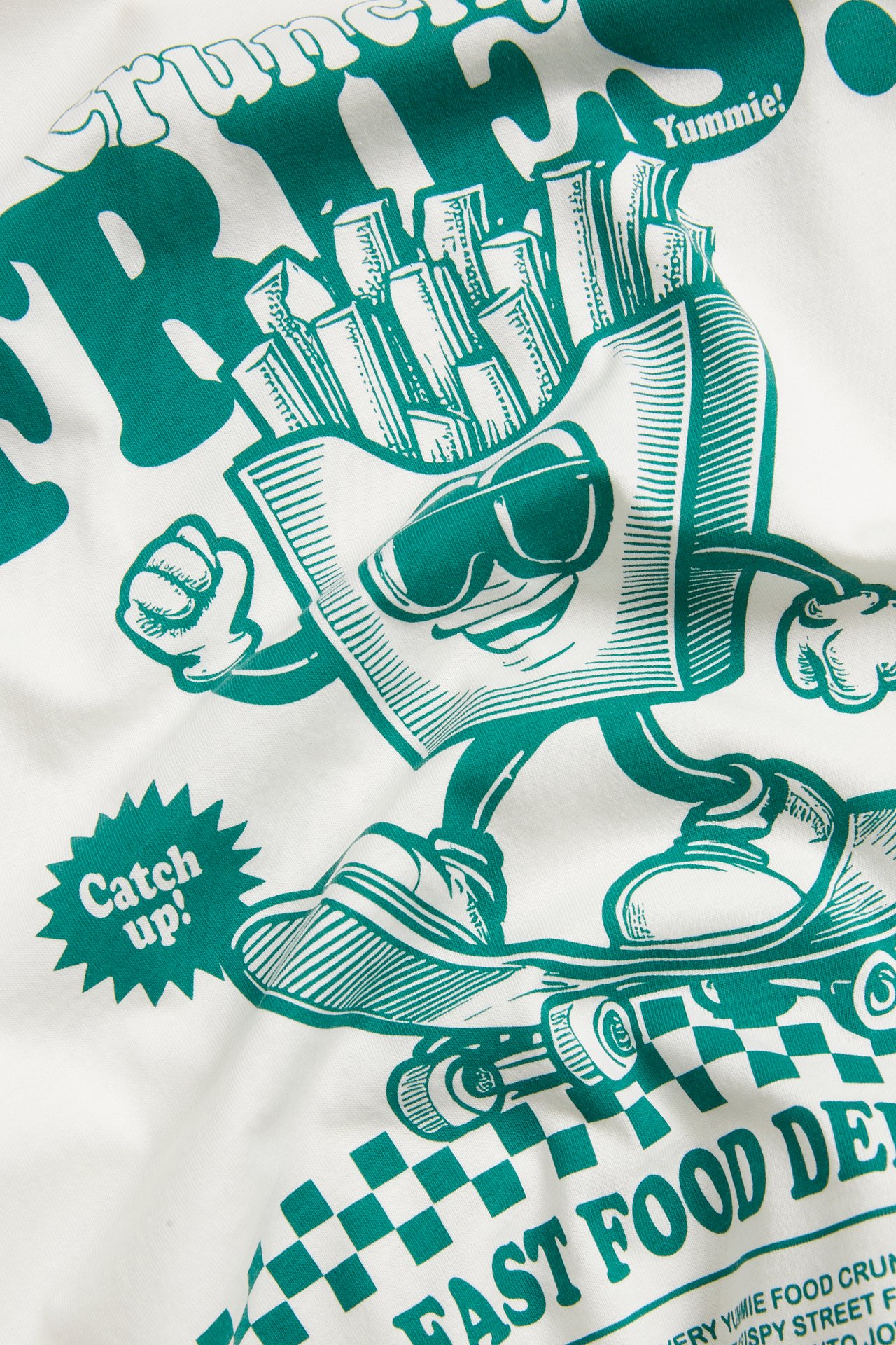 Zbliżenie na bawełnianą koszulkę w kolorze ecru z zielonym nadrukiem postaci frytek na deskorolce.