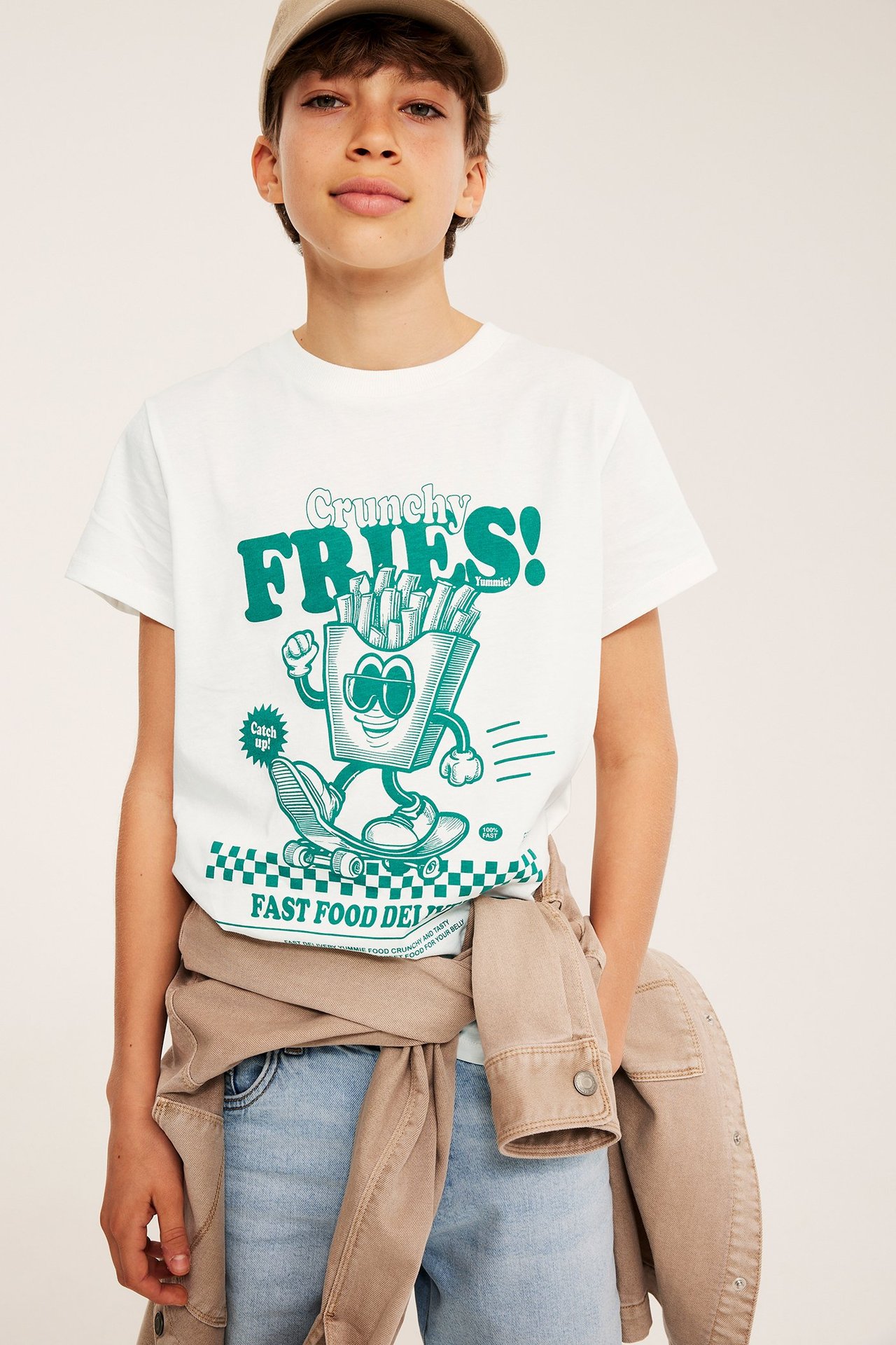 Kid bär offwhite T-shirt med grönt 'Crunchy Fries'-tryck, ljusblå shorts, beige jacka och keps.