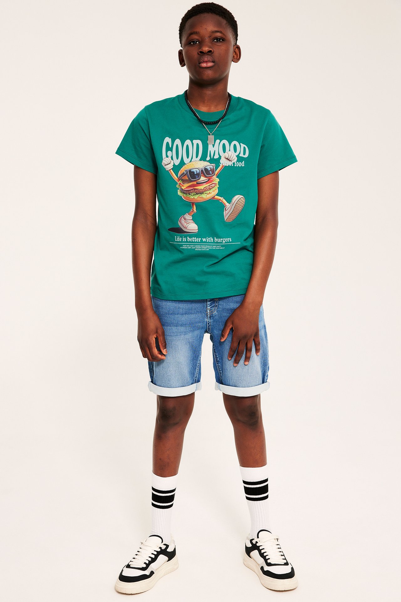 Kid i grön t-shirt med hamburgartryck, ljusblå denimshorts, vita randiga strumpor och svarta sneakers.