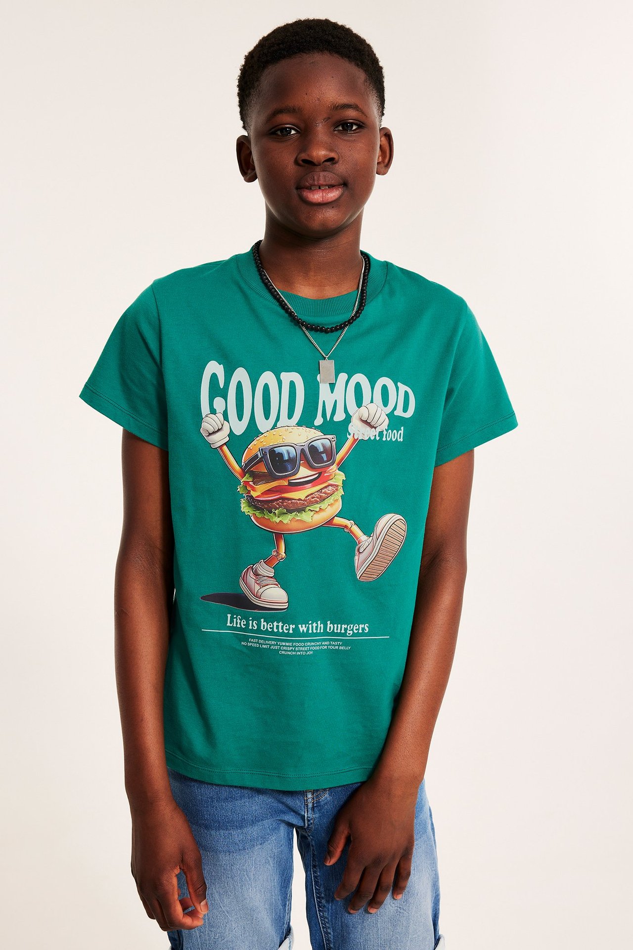 Framifrån: Kid bär grön kortärmad T-shirt med burgartryck, blå jeansshorts och ett pärlhalsband.