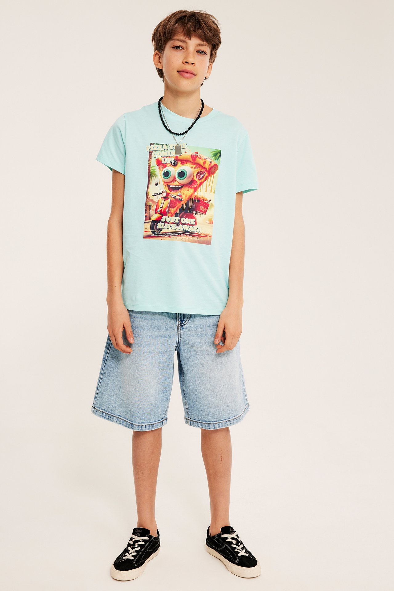 Framifrån: kille i ljus turkos T-shirt med färgglatt tryck, ljusa jeansshorts och svarta sneakers.