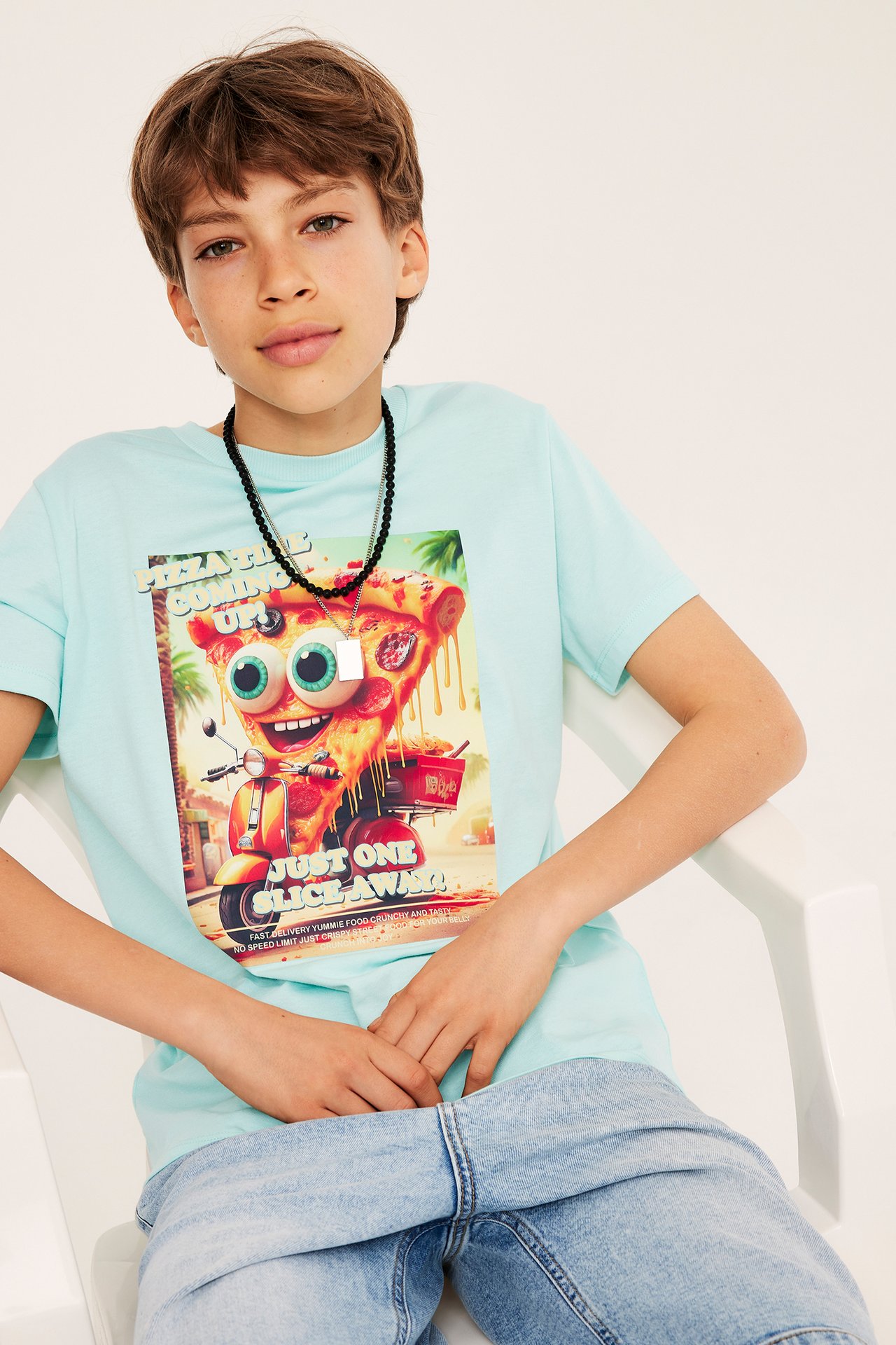 Forfra: gutt i lys turkis bomulls-T-skjorte med pizzatrykk, lyse denimshorts og svart perlekjede.