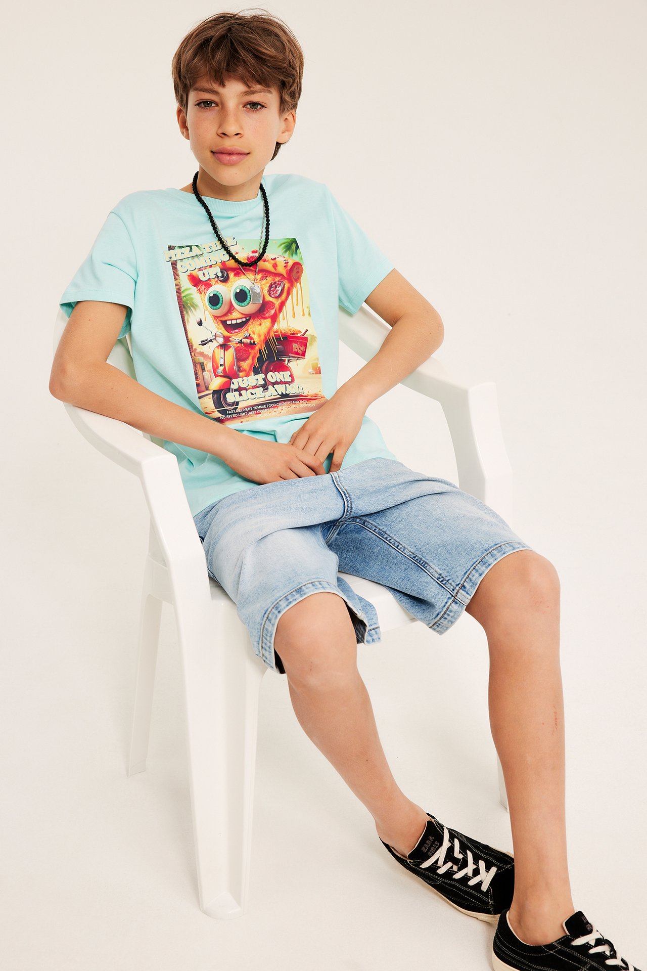 Framsida: Kid bär ljus turkos bomulls-t-shirt med pizzatryck, ljusblå denimshorts och svarta sneakers.