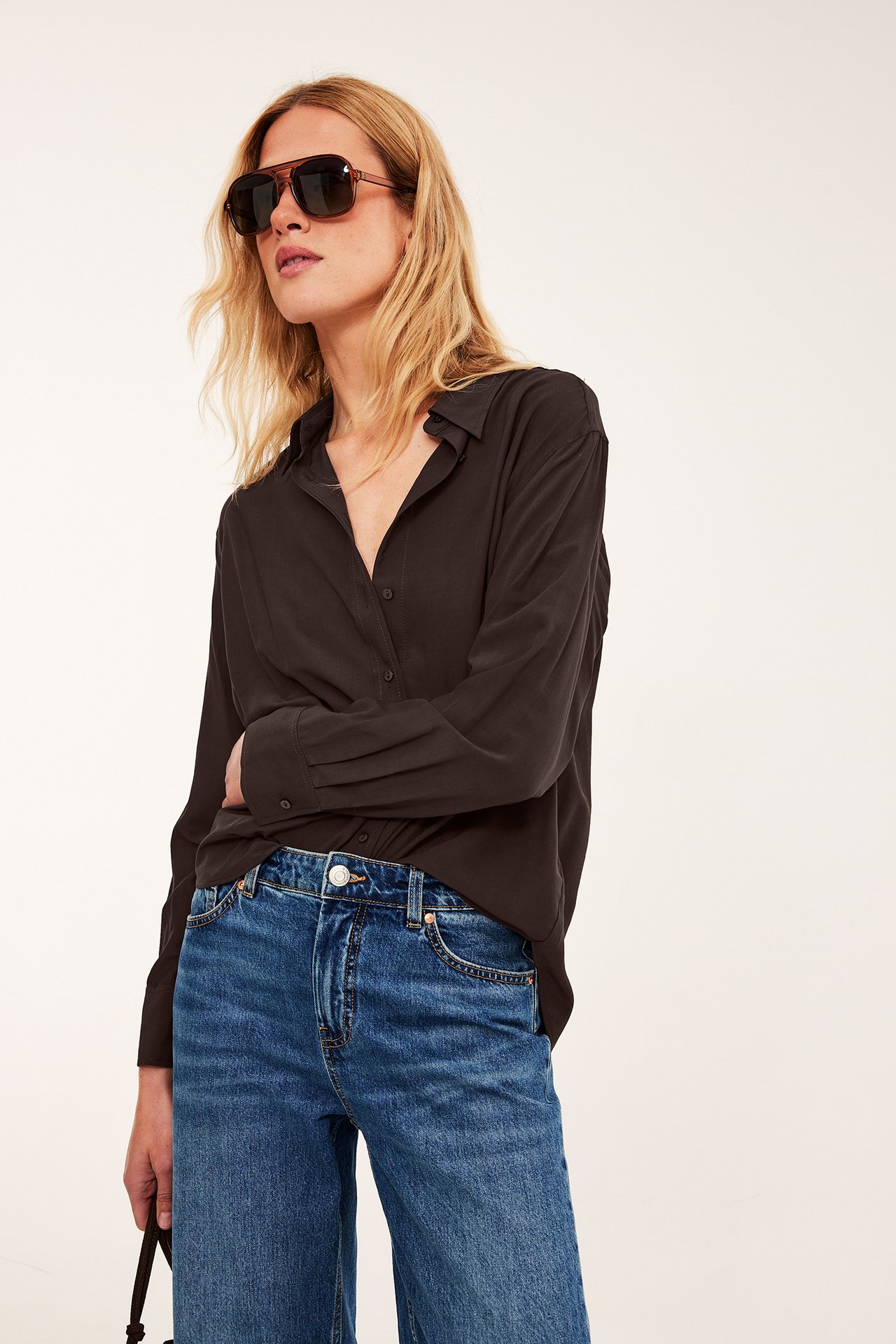 Framifrån: kvinna i mörkbrun långärmad skjorta, blå vida jeans och bruna solglasögon.