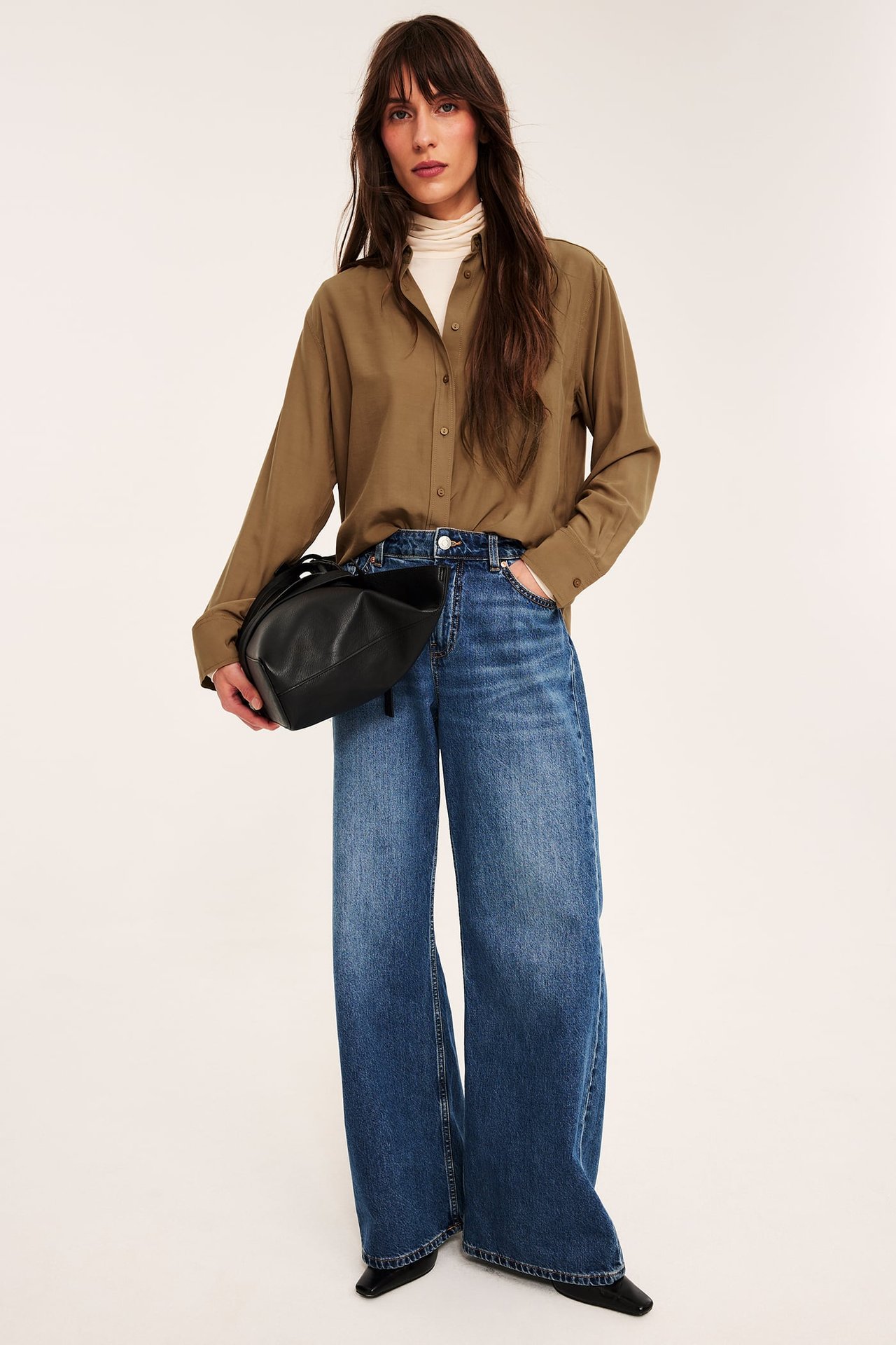 Kvinna i olivgrön skjorta, beige polotröja, blå vida jeans och håller svart handväska.