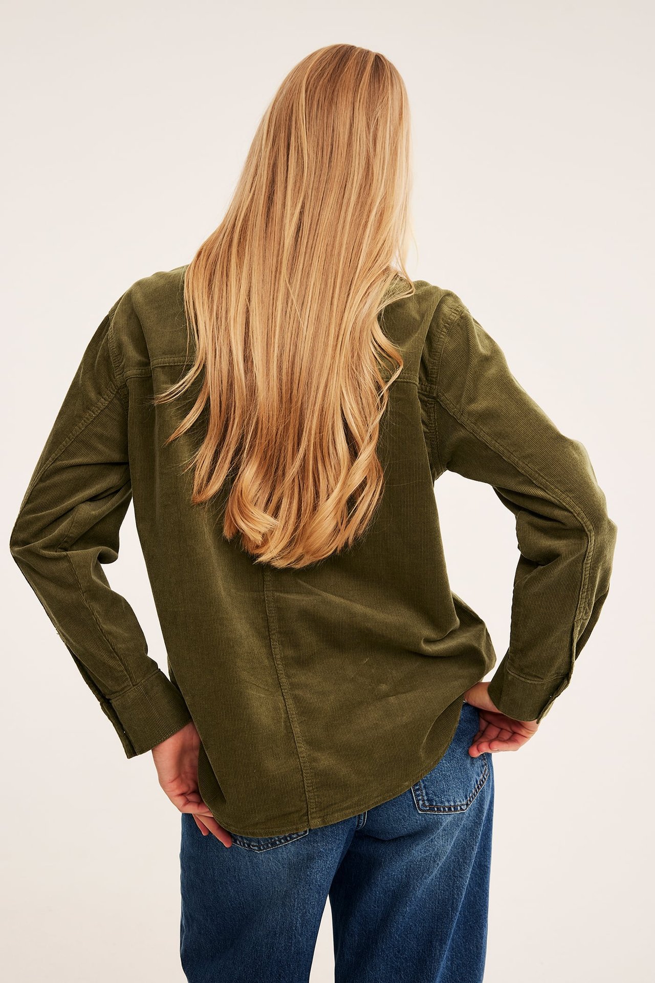 Ryggvisning av kvinnelig modell i mørkegrønn kordfløyelsskjorte og blå jeans.