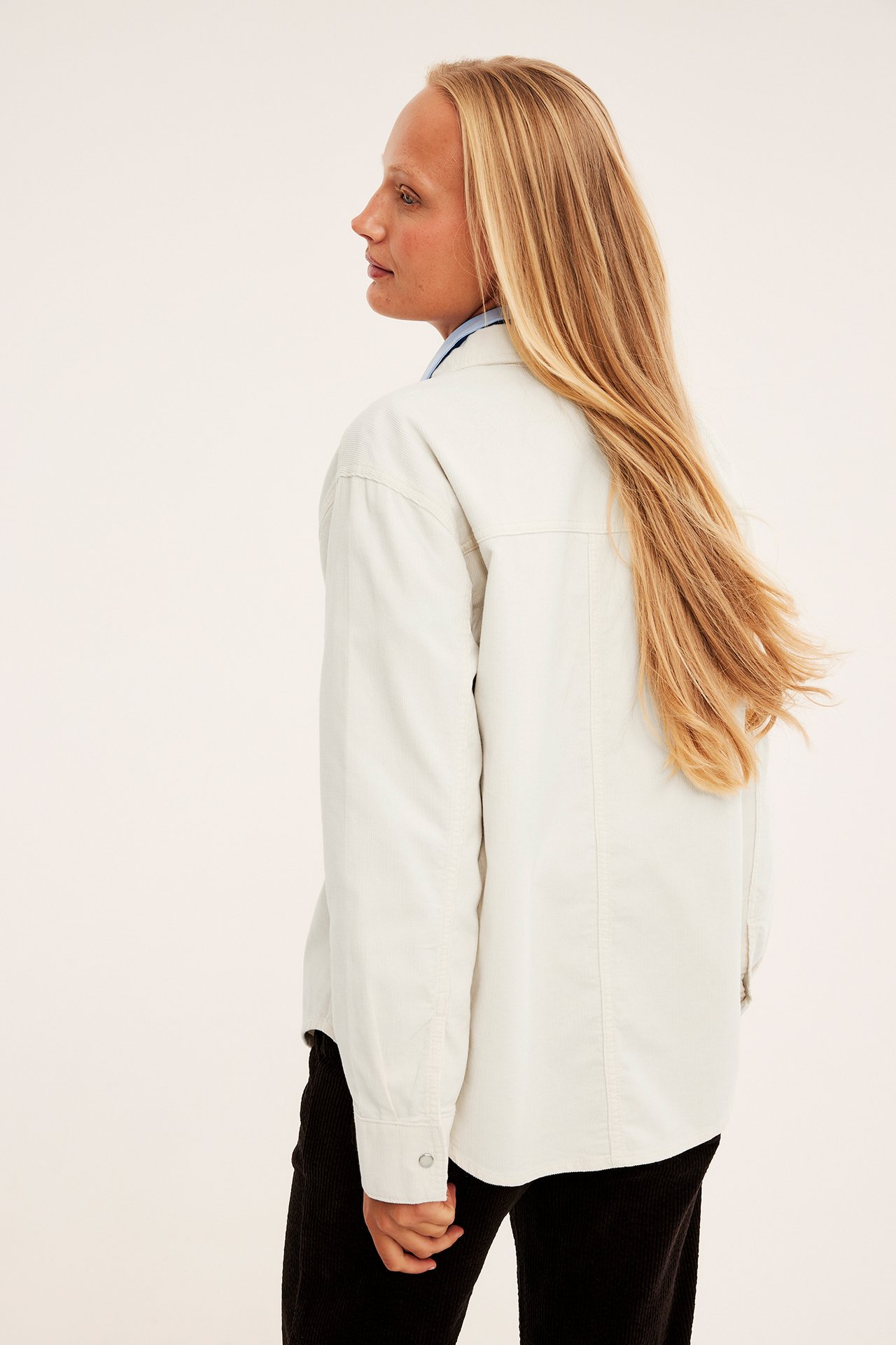 Sidevisning av kvinne i lys beige oversized skjortejakke over blå skjorte og svarte ribbestrikkede bukser.