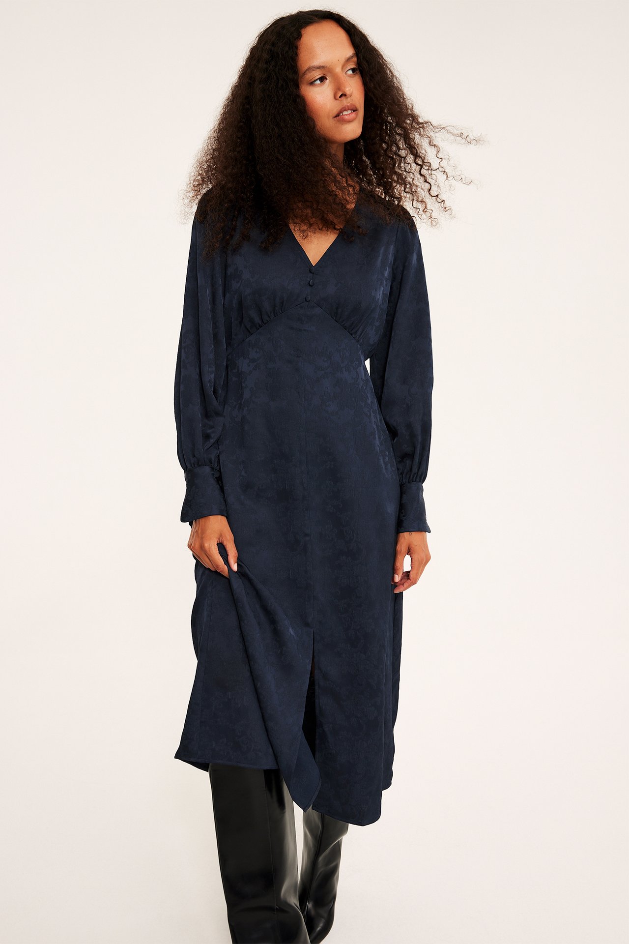 Frontvy av kvinnlig modell i marinblå jacquard satinklänning med långa ärmar och svarta stövlar.