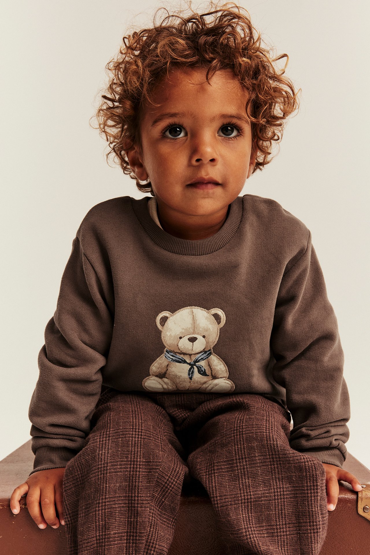 Frontvy av ett barn i en brun sweatshirt med teddybjörnstryck och bruna rutiga byxor.