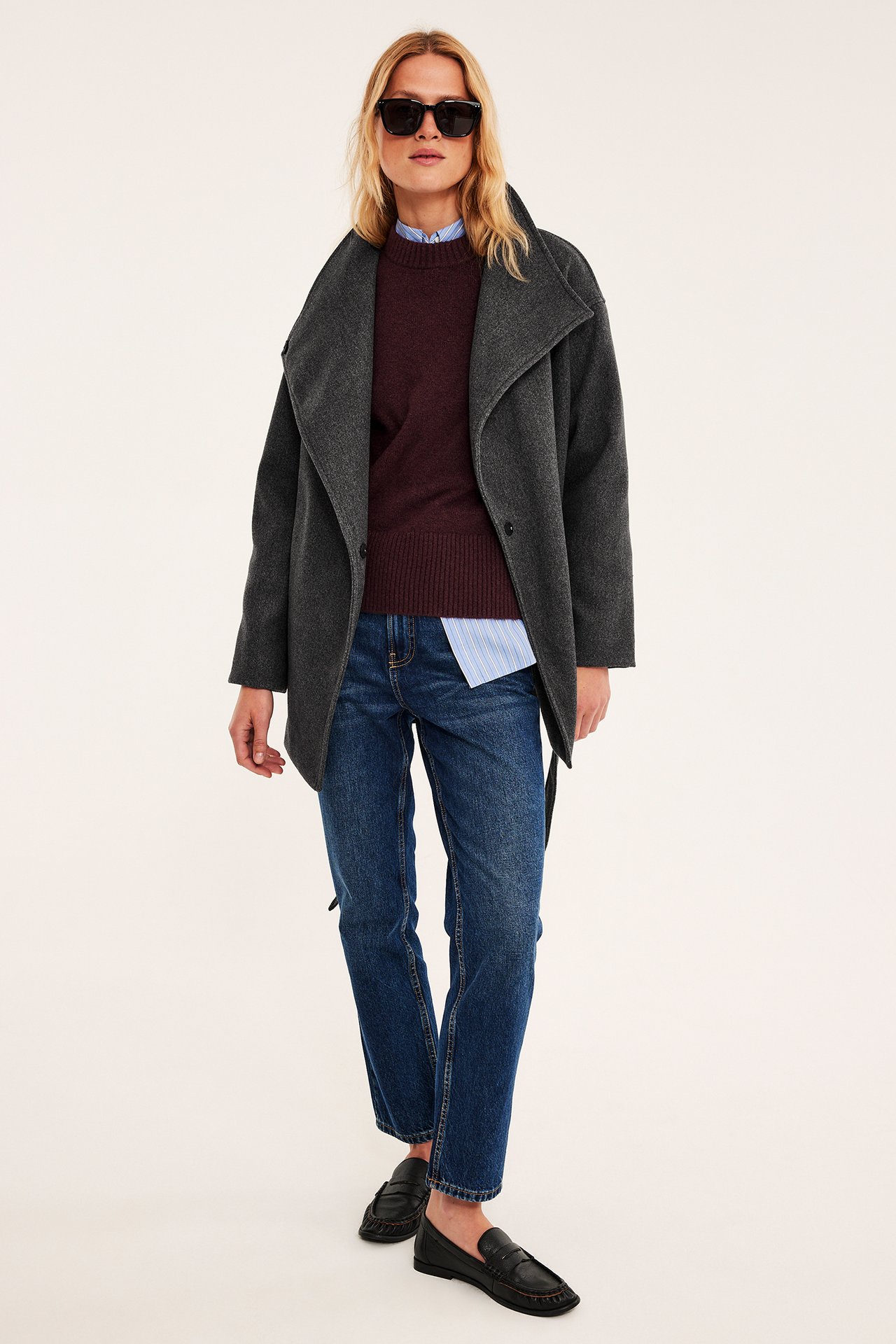Kvinnas mörkgrå jacka över vinröd tröja, randig skjorta, blå jeans och svarta loafers, framifrån.